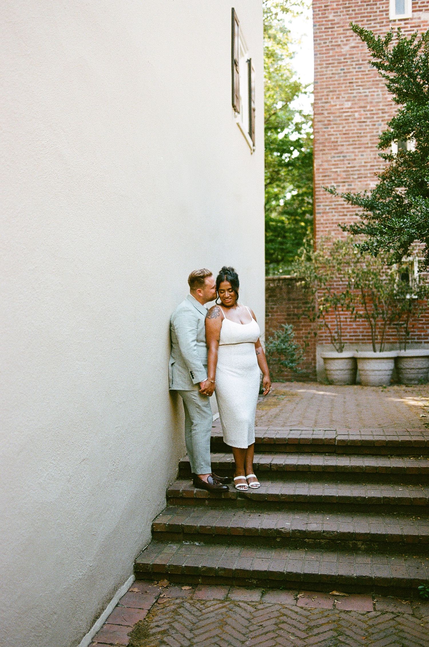 A casual couple stands close together on brick steps between white and red brick walls.