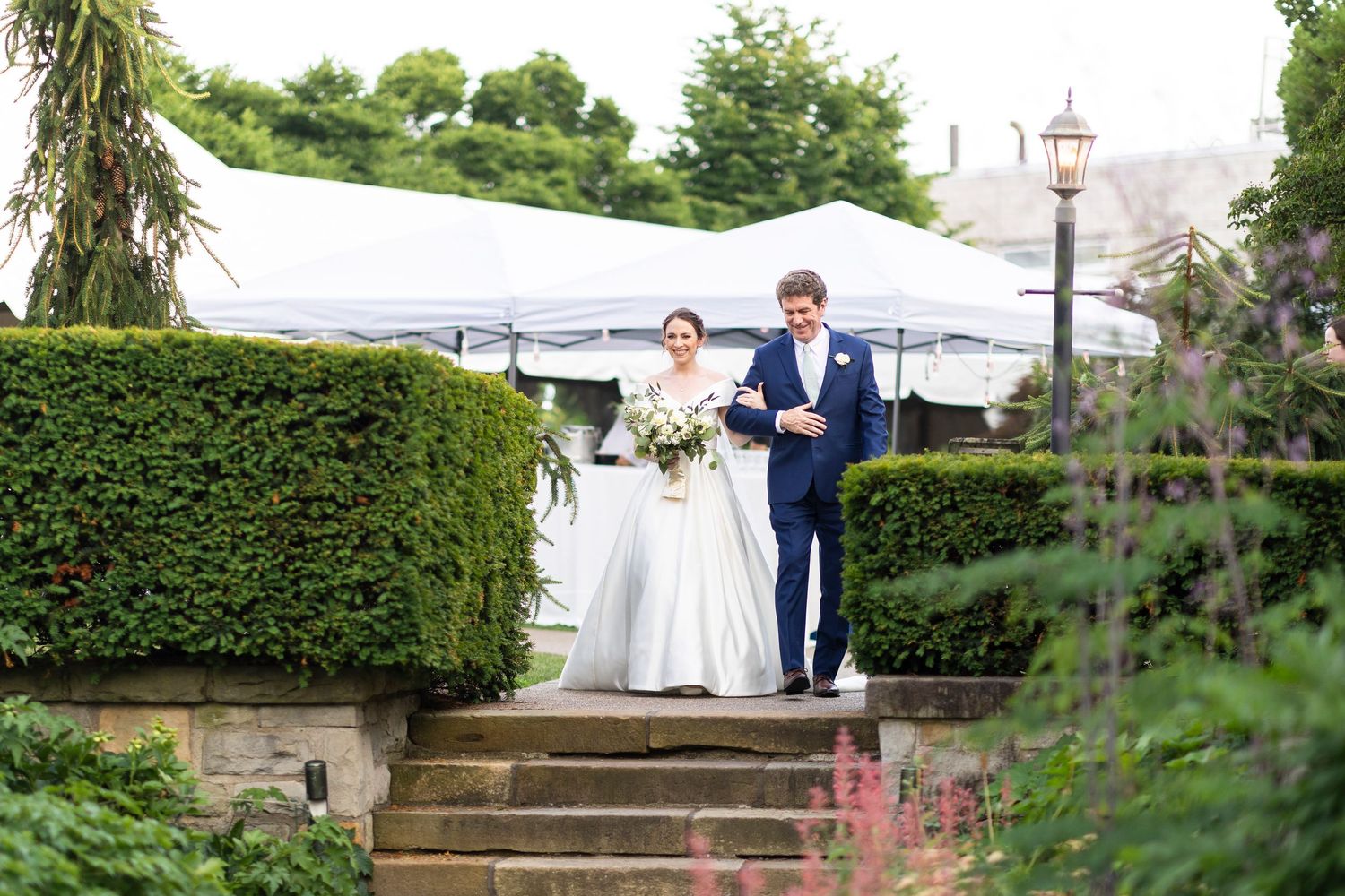Wedding party members walk along garden pathways with decorative hedges and stone steps.