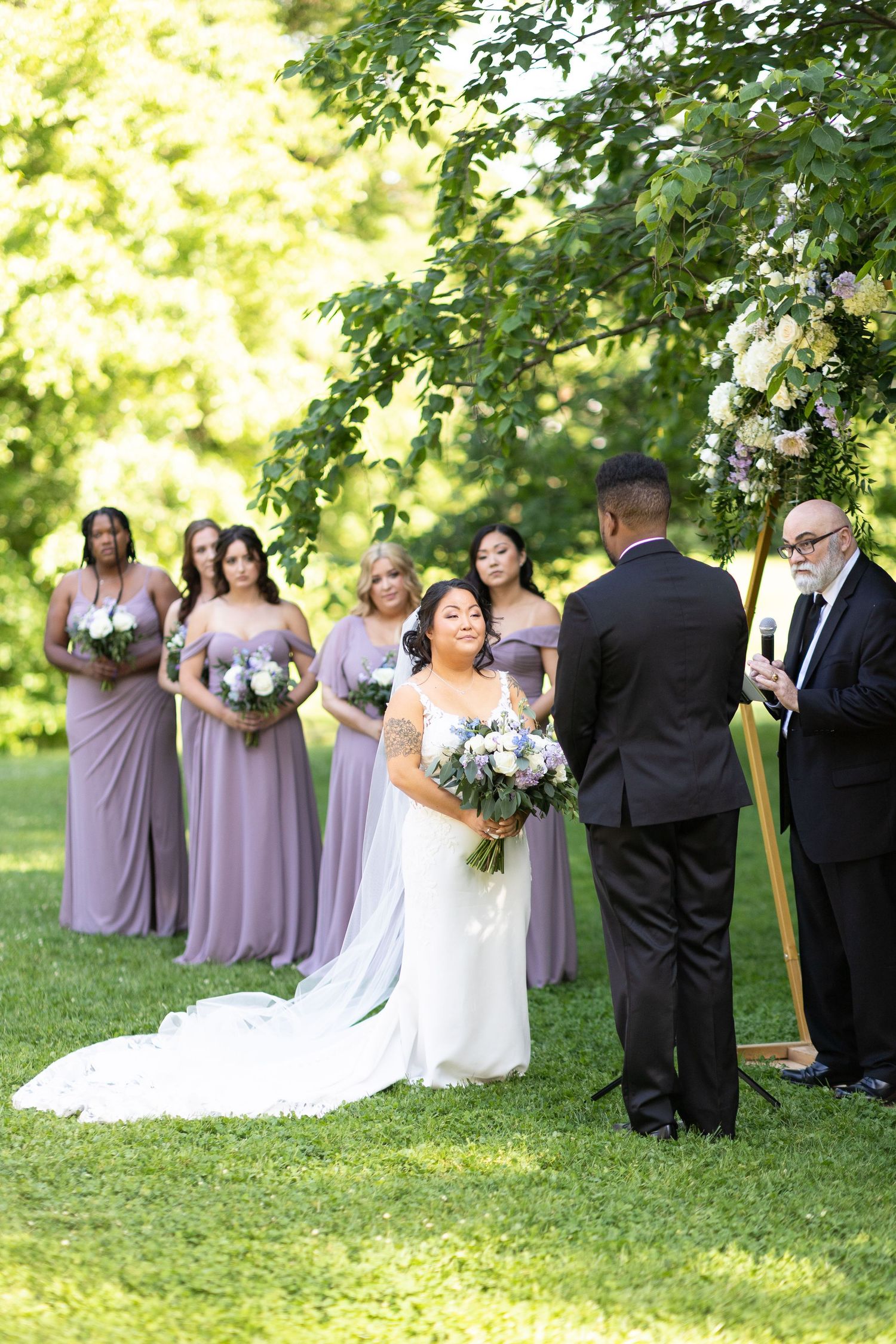 Wedding party in purple dresses and dark suits poses on sunny grass under trees.