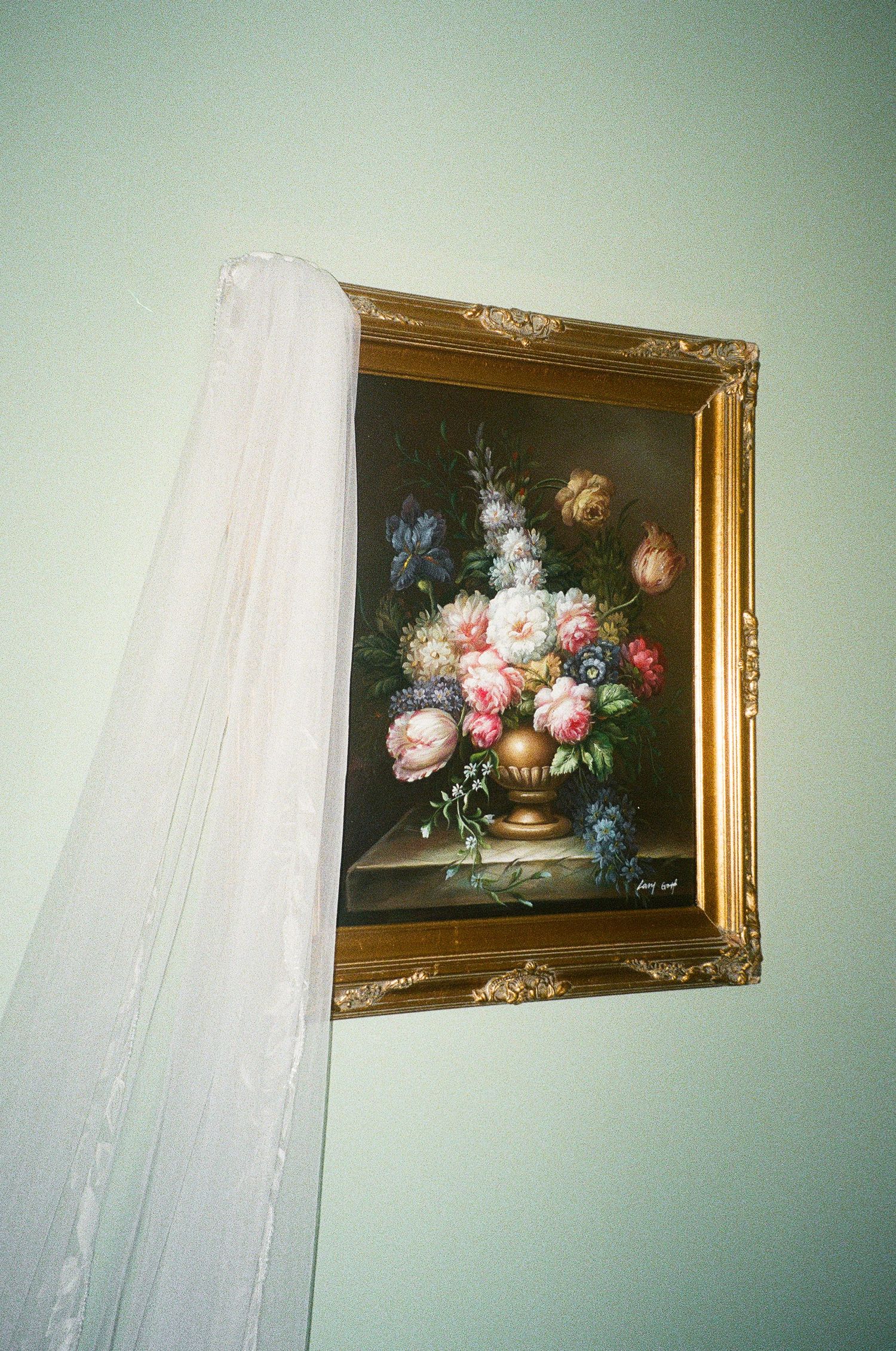 Gold ornate framed floral oil painting with roses and mixed flowers in a vase, hanging on a mint green wall.