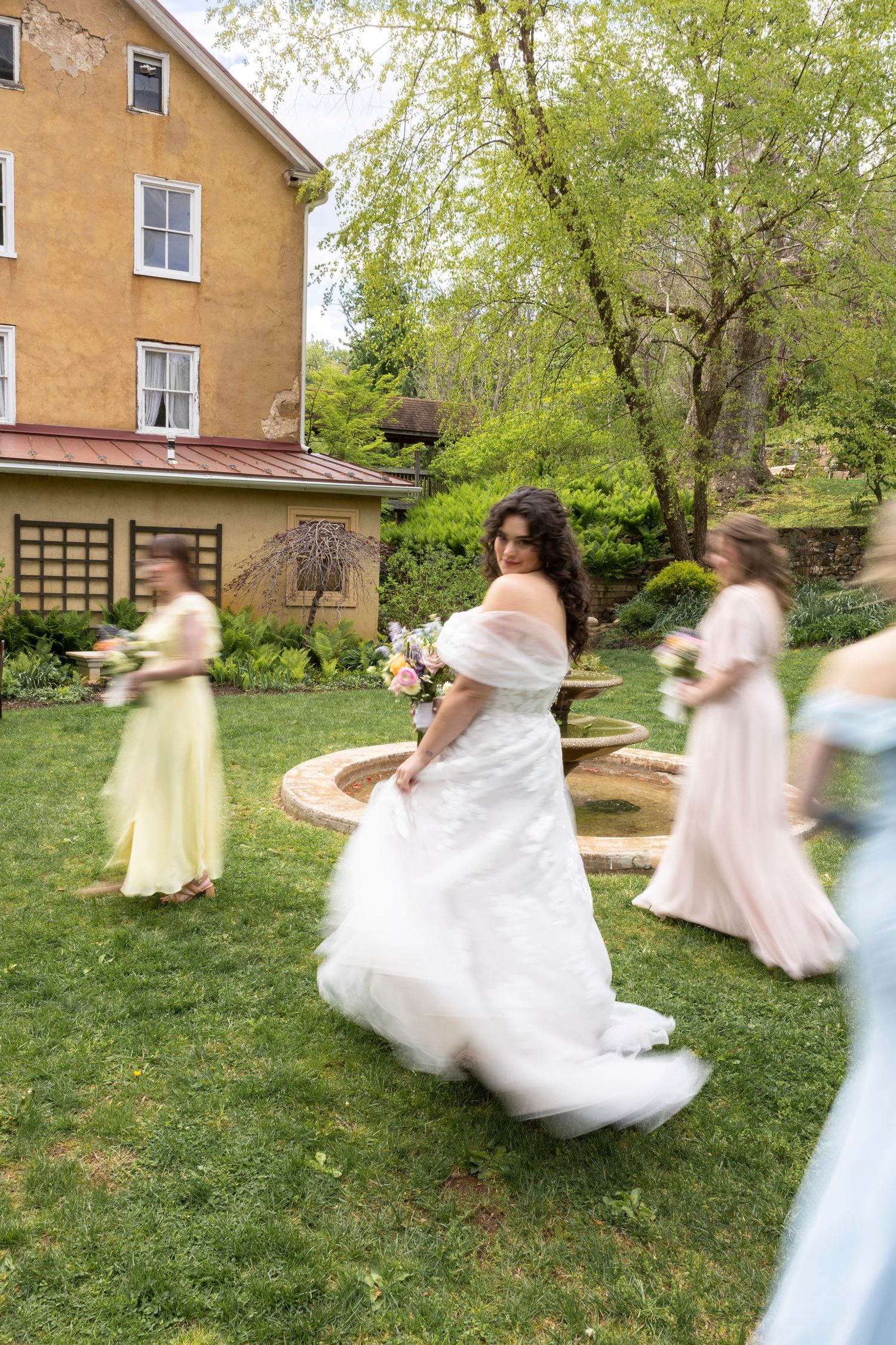 Bride in white off-shoulder gown twirls with bridesmaids in pastel dresses on green lawn beside historic yellow building.