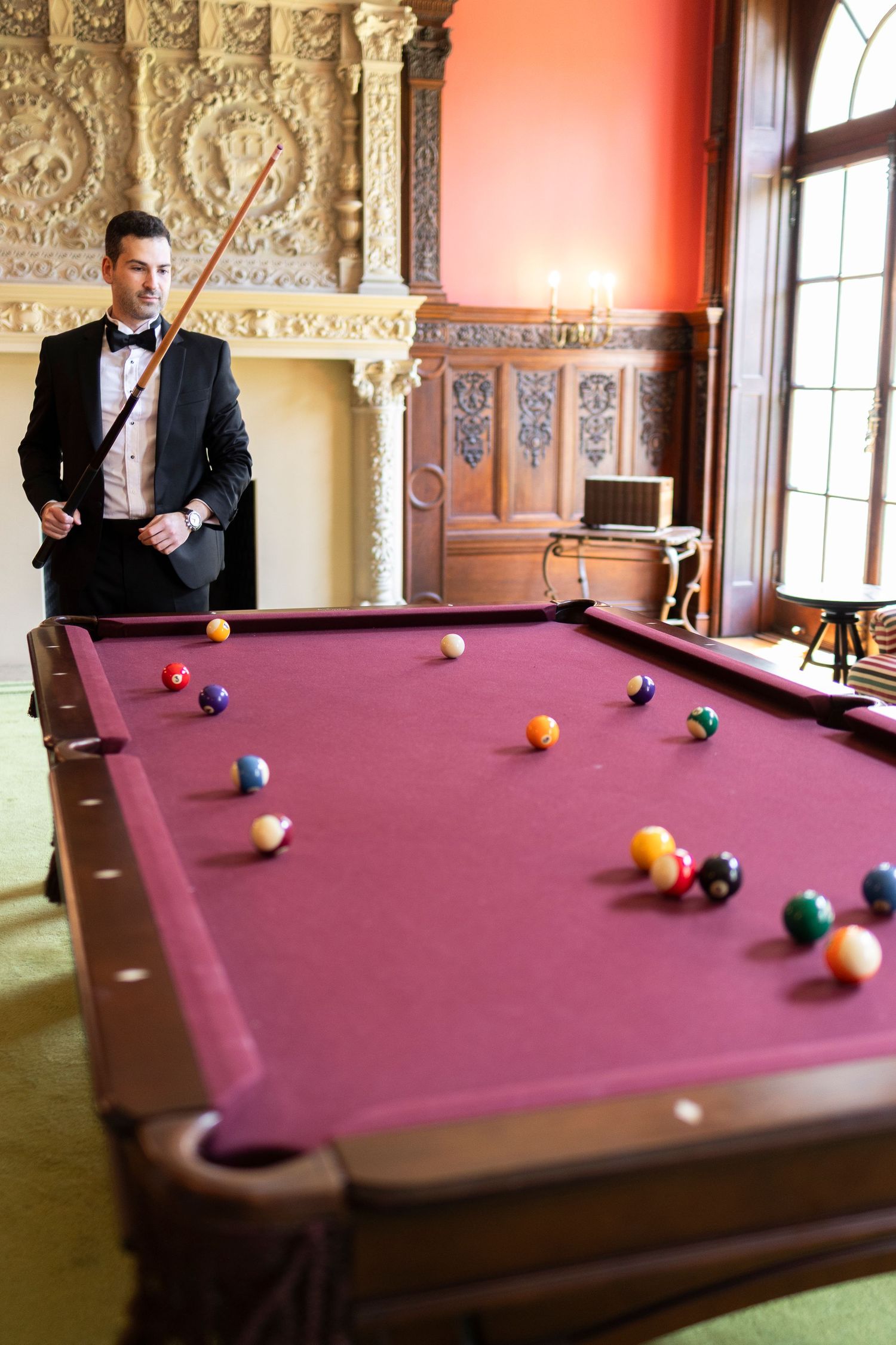 Elegant man in tuxedo holding billiard cue beside a pool table with colorful balls in a luxurious room.