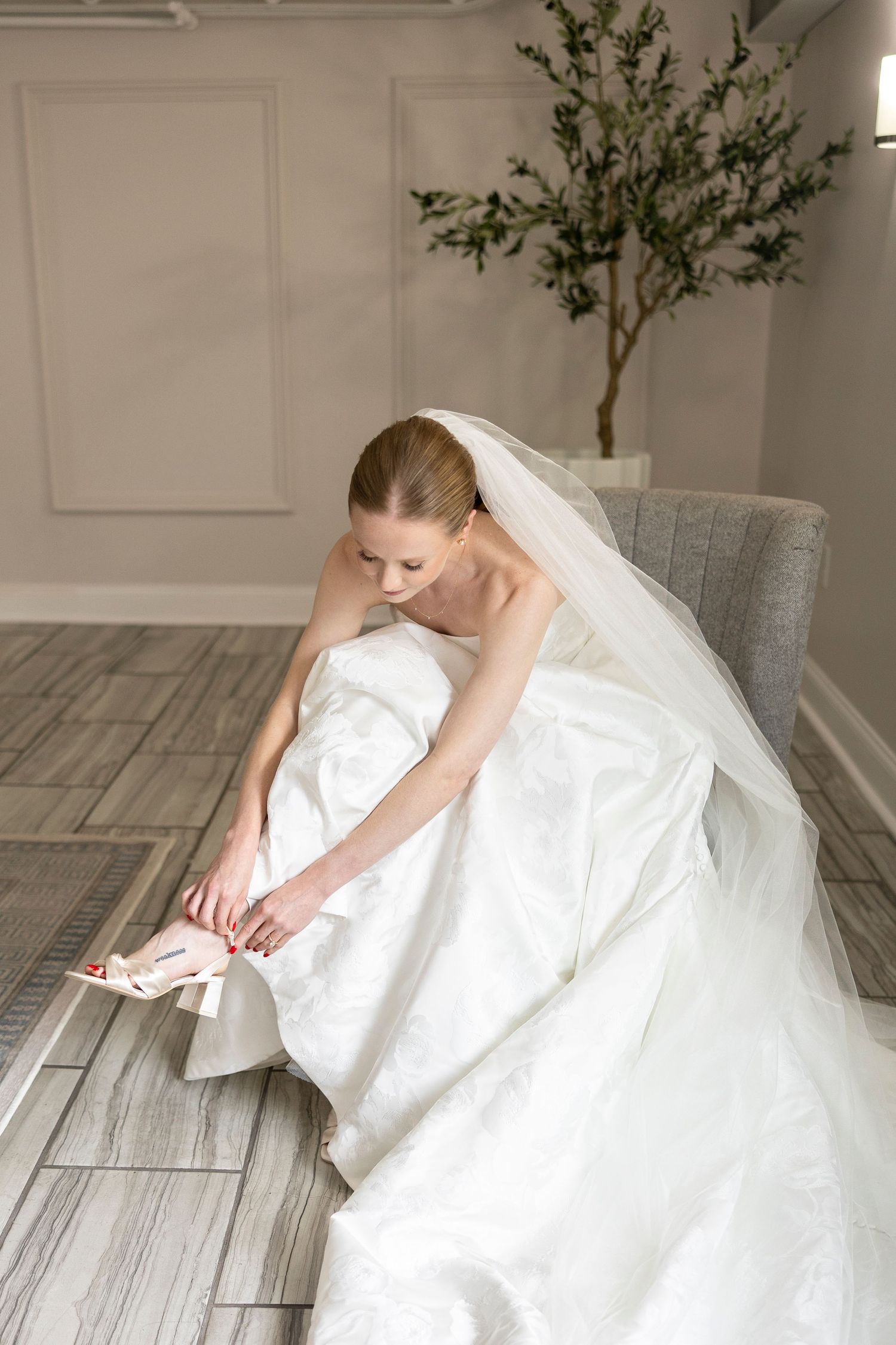 Bride in white gown sitting elegantly on grey chair with flowing veil.