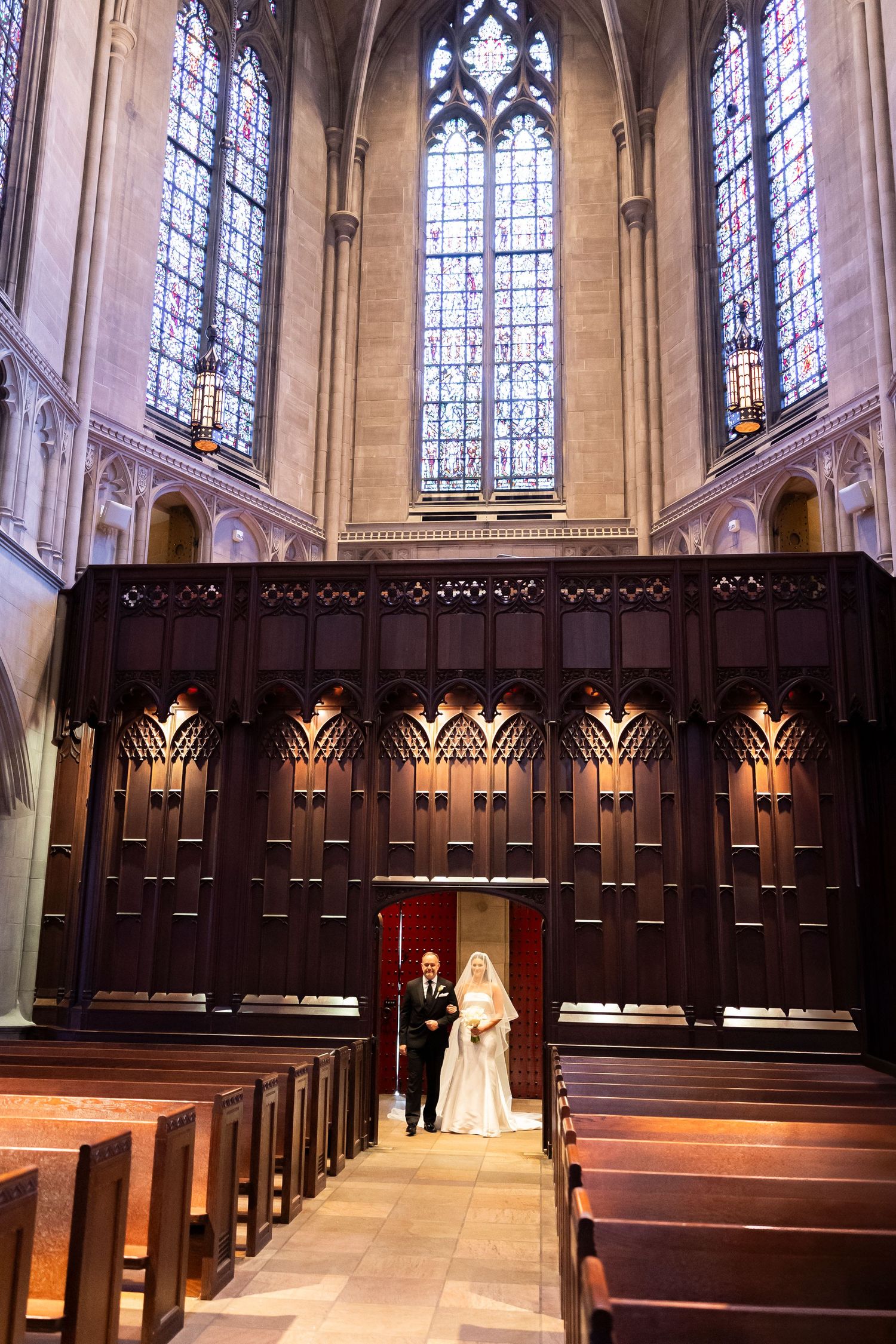 Bride and groom stand at the altar of a grand cathedral with ornate wooden panels and stained glass windows.