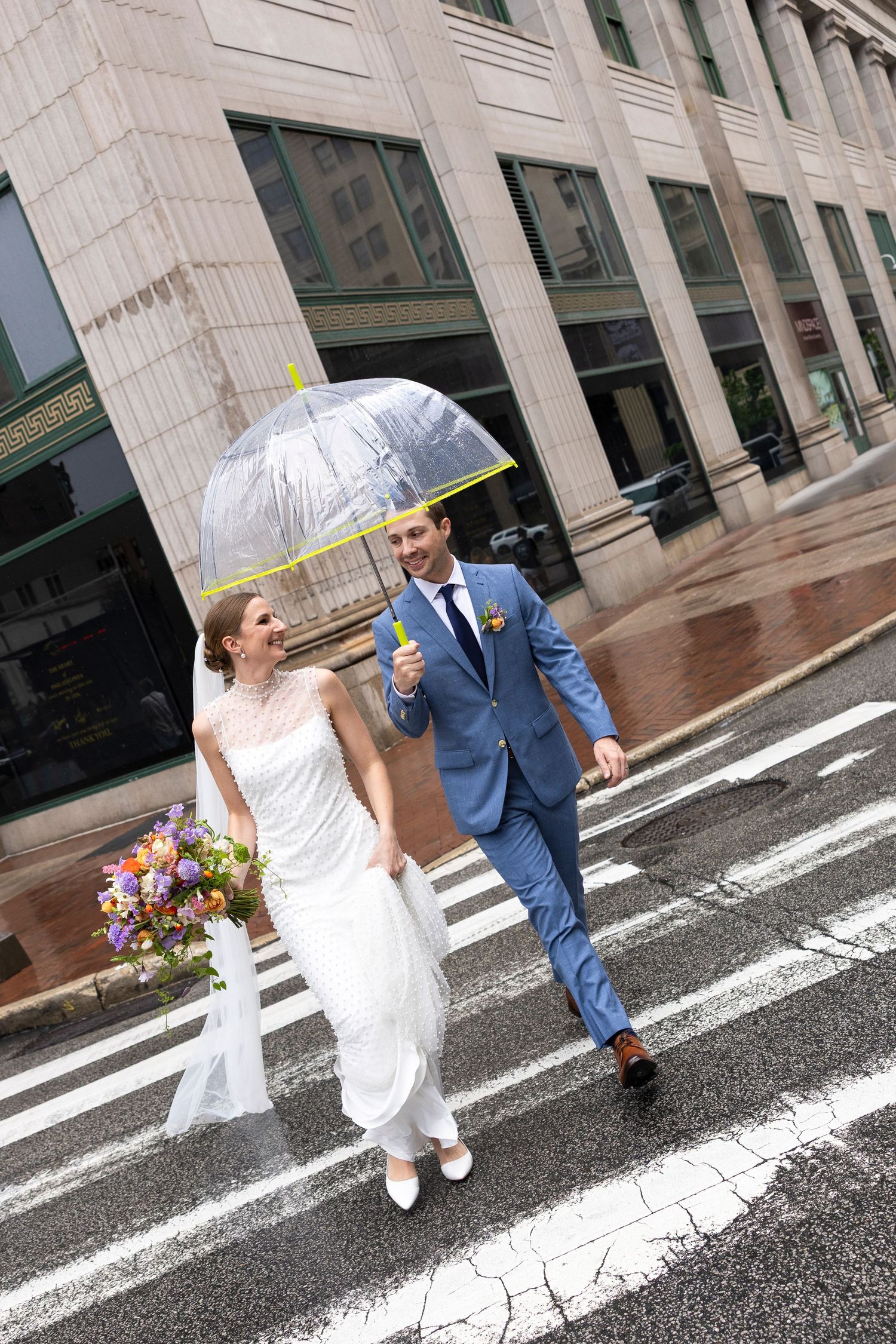 Bride and groom crossing a wet street in the rain, holding a clear umbrella and colorful bouquet, dressed in wedding attire.