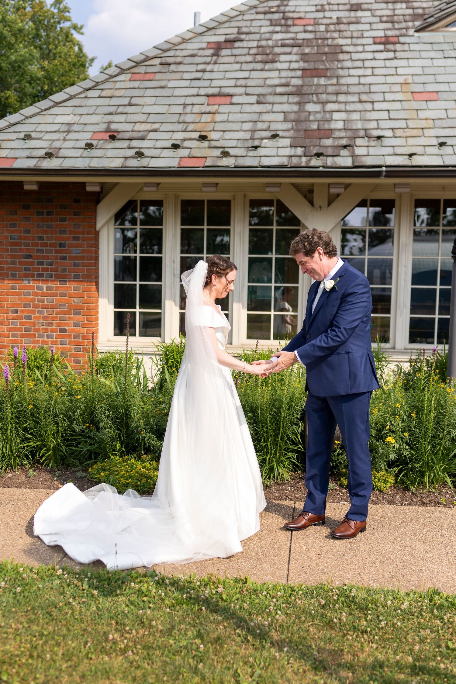 A couple in wedding attire playfully interact on a garden path near a brick building.