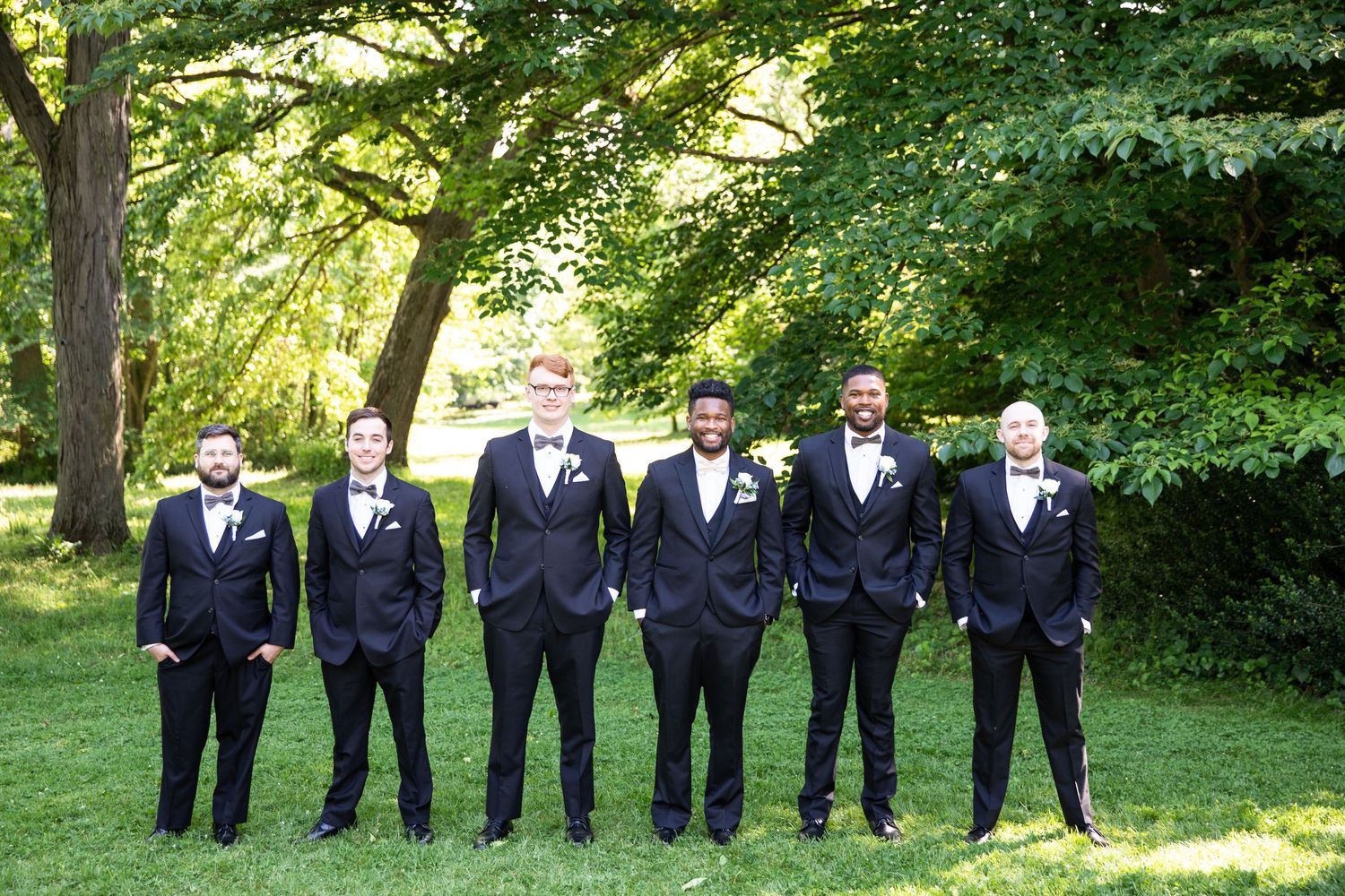 Groomsmen in black suits and bowties pose in line on grass under trees.