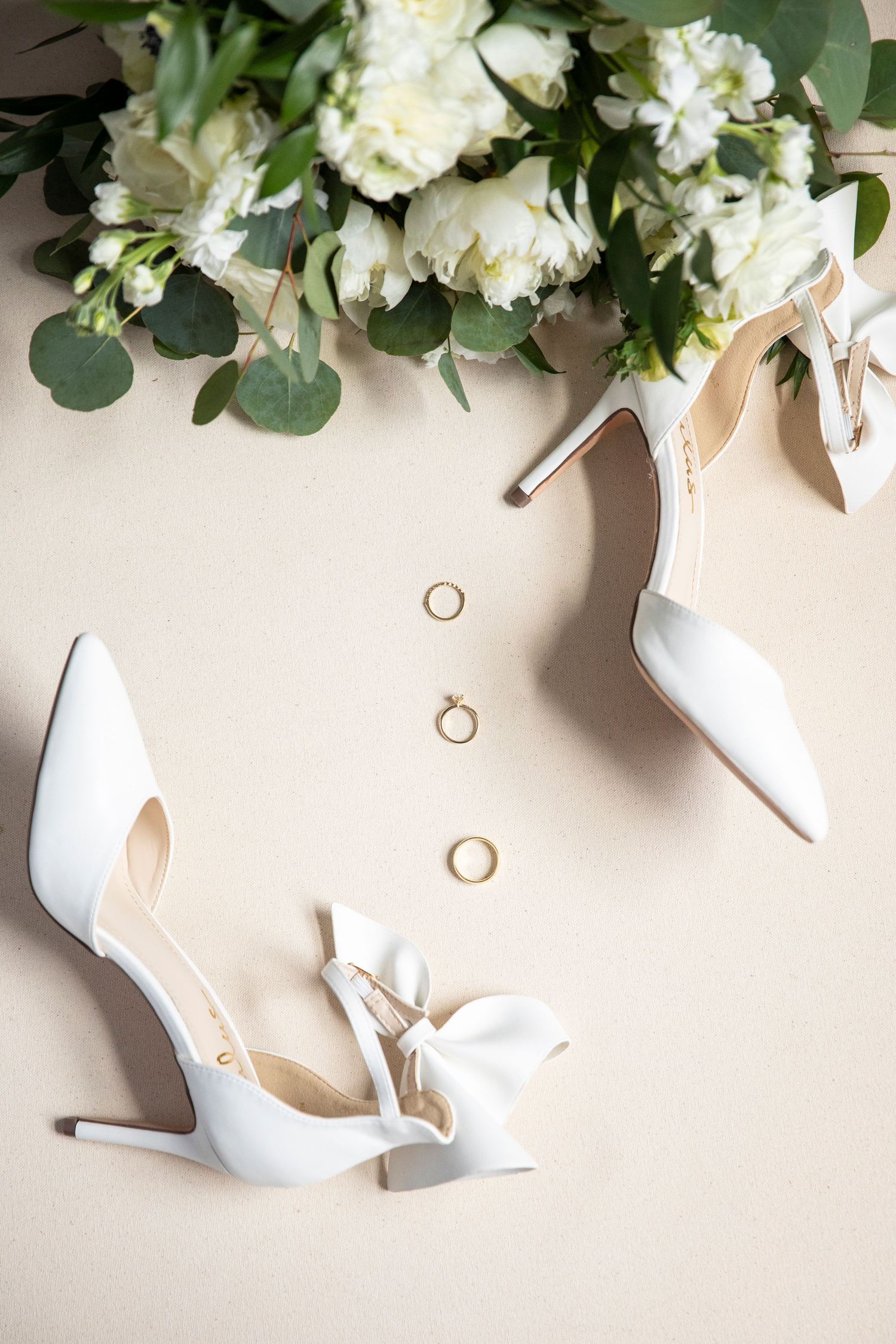 White bridal heels with wedding rings and floral arrangement styled on beige background.