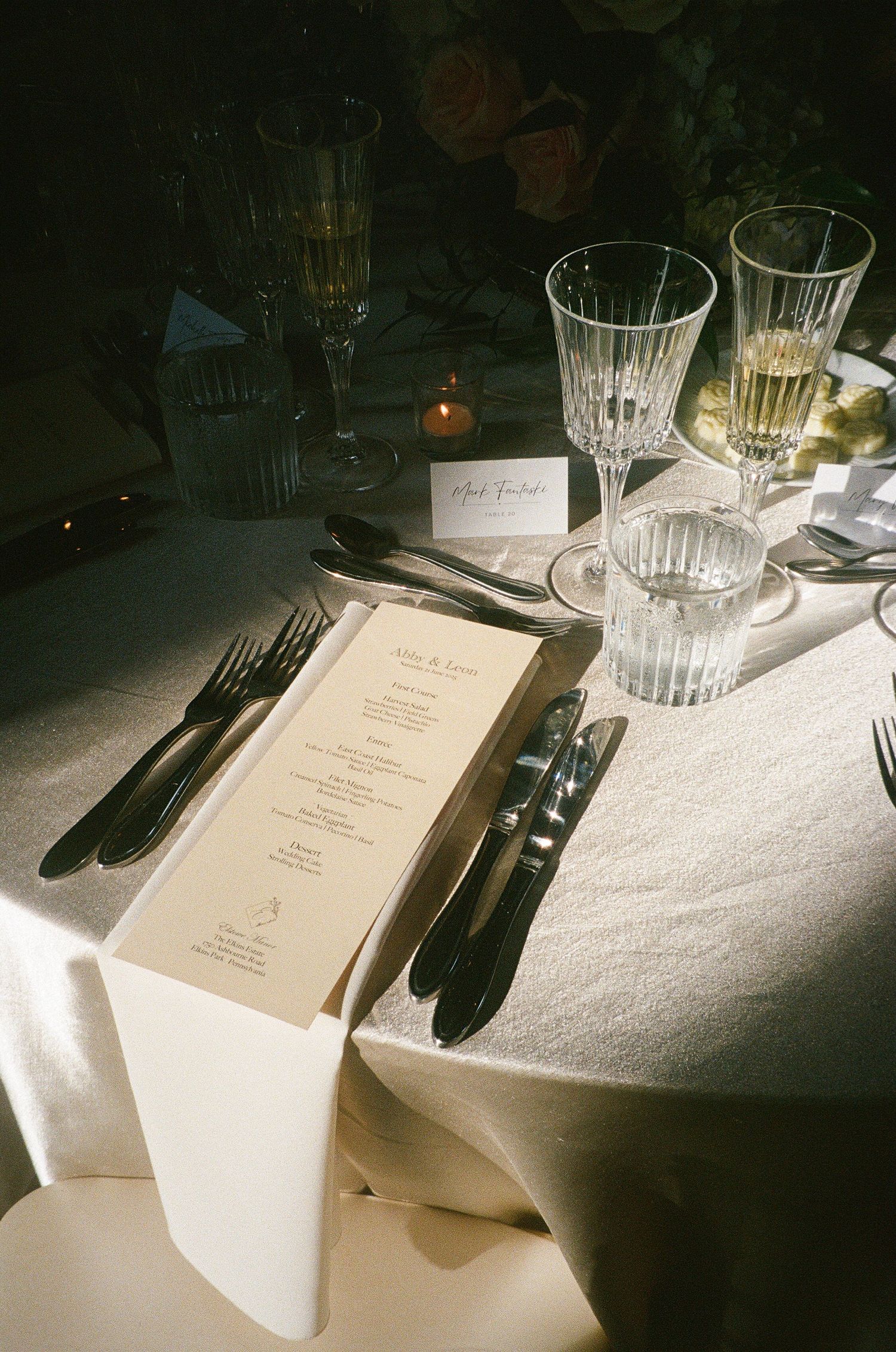 Formal place settings with crystal glasses and menus on white tablecloths in dim lighting.