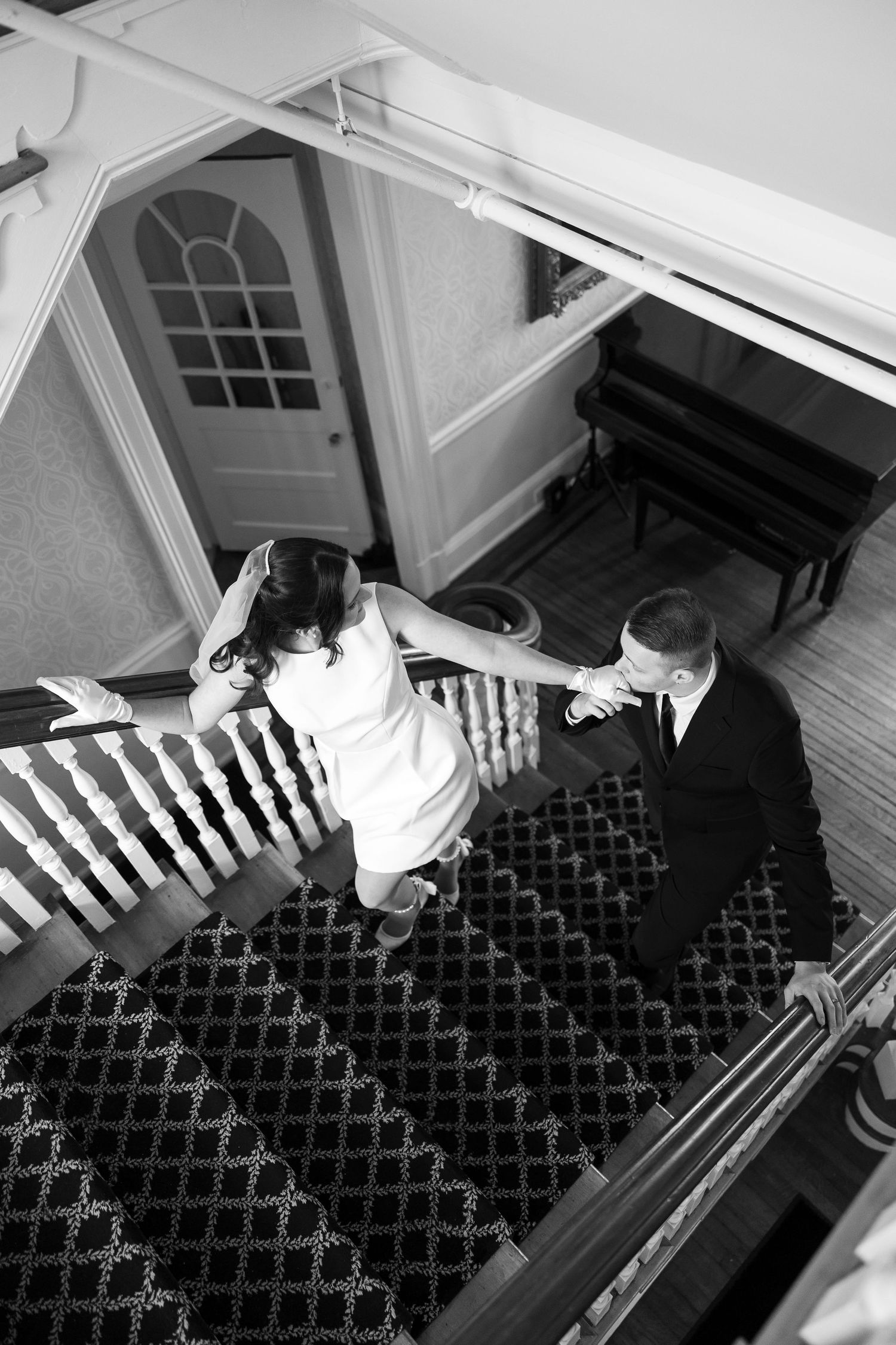 A couple descends an elegant staircase in formal attire, viewed from above.