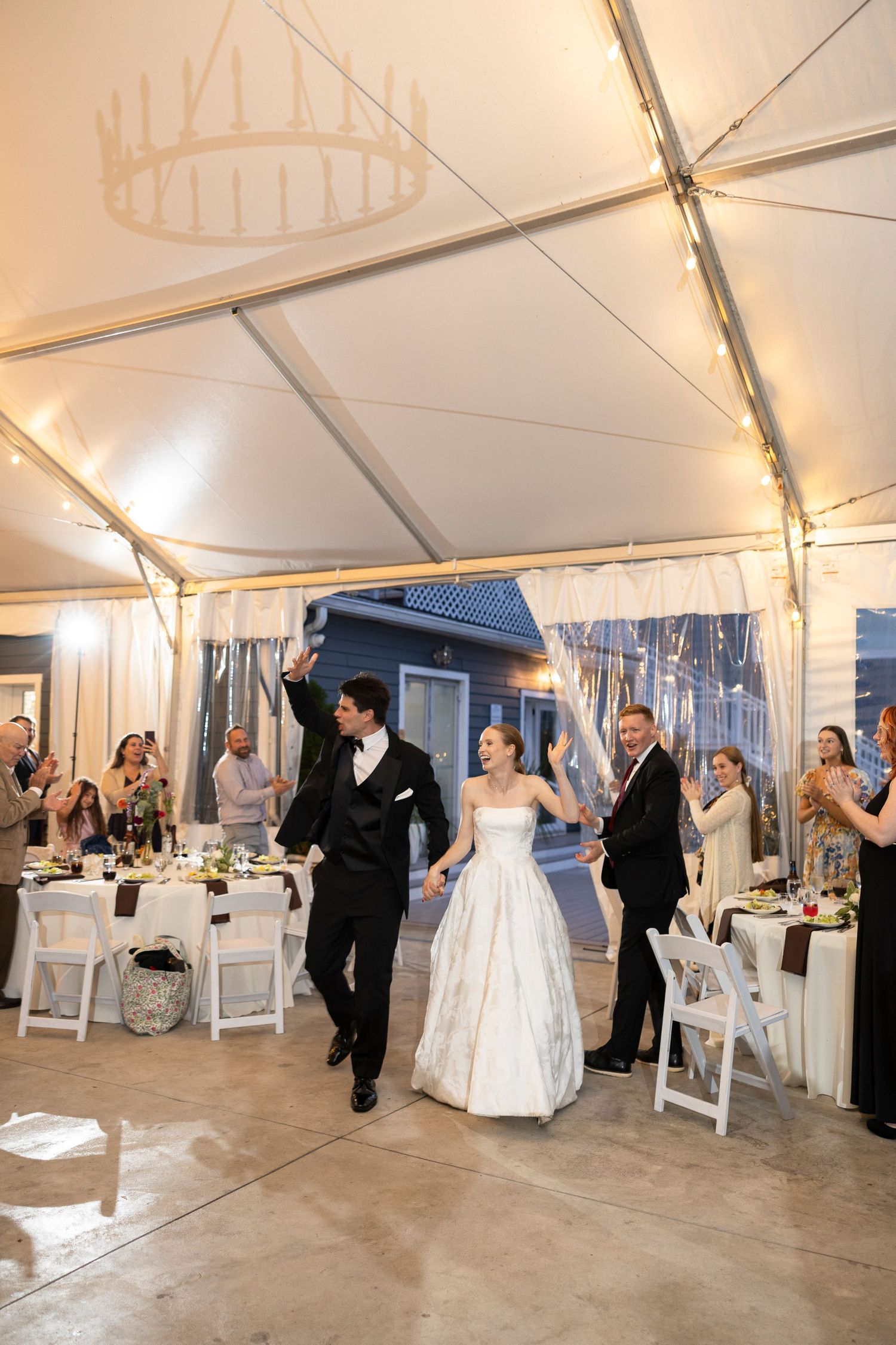 Wedding reception in progress under white tent with string lights and white folding chairs.