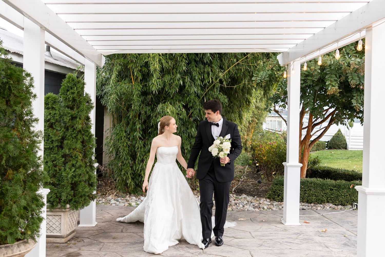 A wedding couple walks under an elegant white pergola in a garden setting.