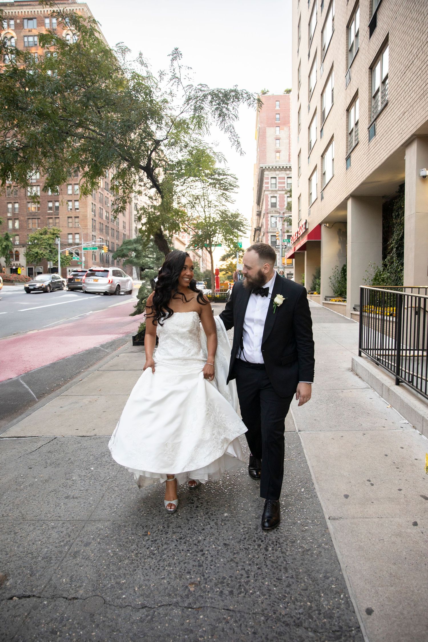 Wedding couple walking along urban sidewalk with apartment buildings in background.