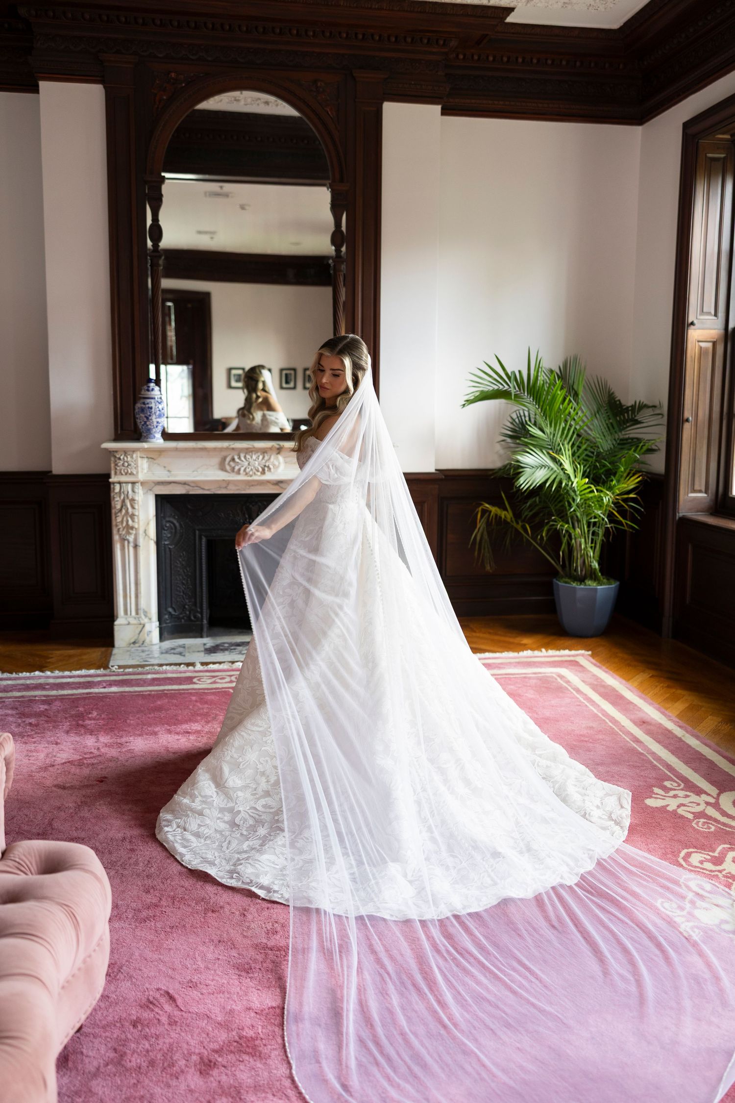 Wedding dress with long train spreads across pink carpet in an elegant room with dark wood trim.