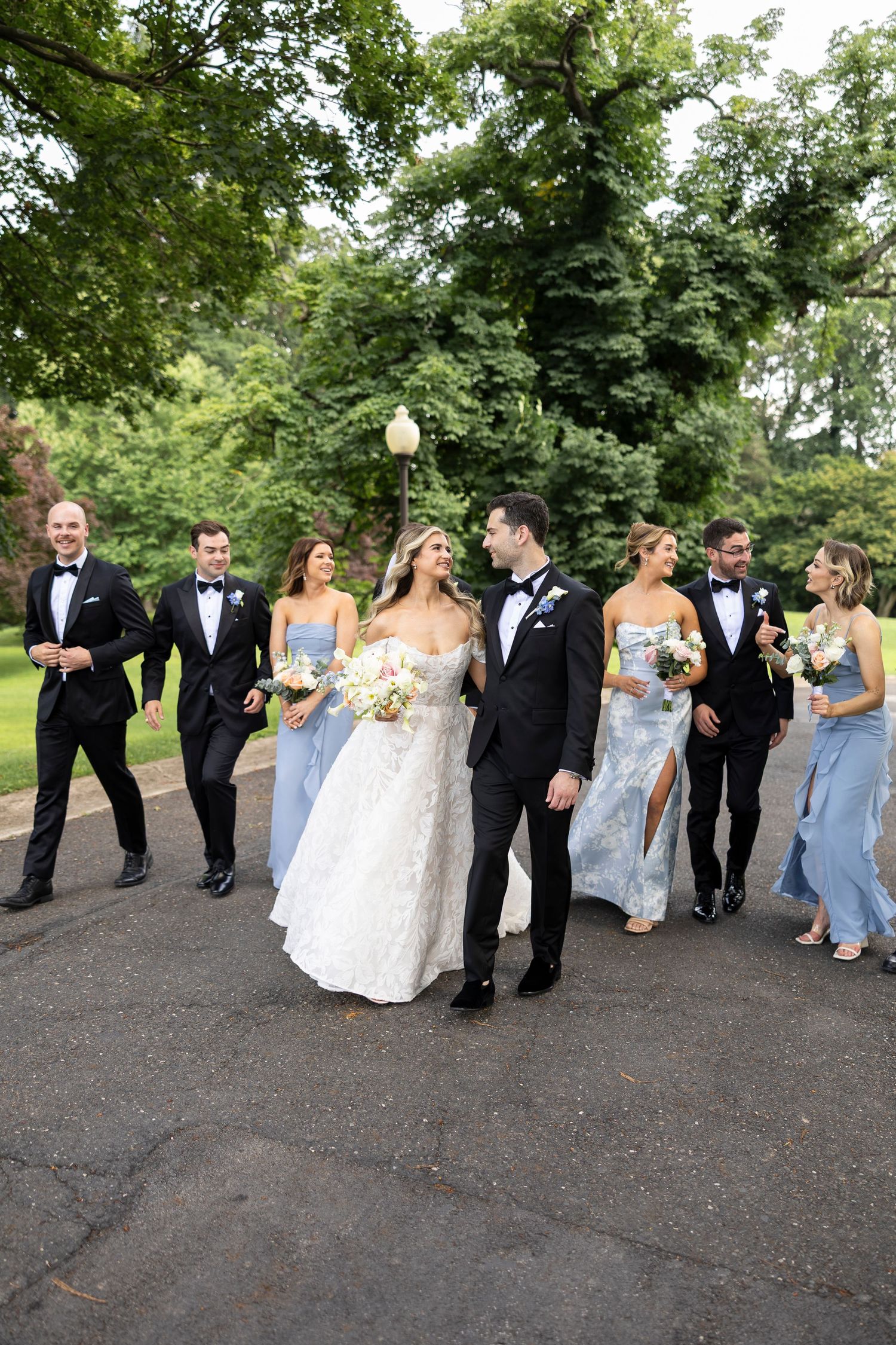 Wedding party walking and laughing together on a path surrounded by lush green trees.