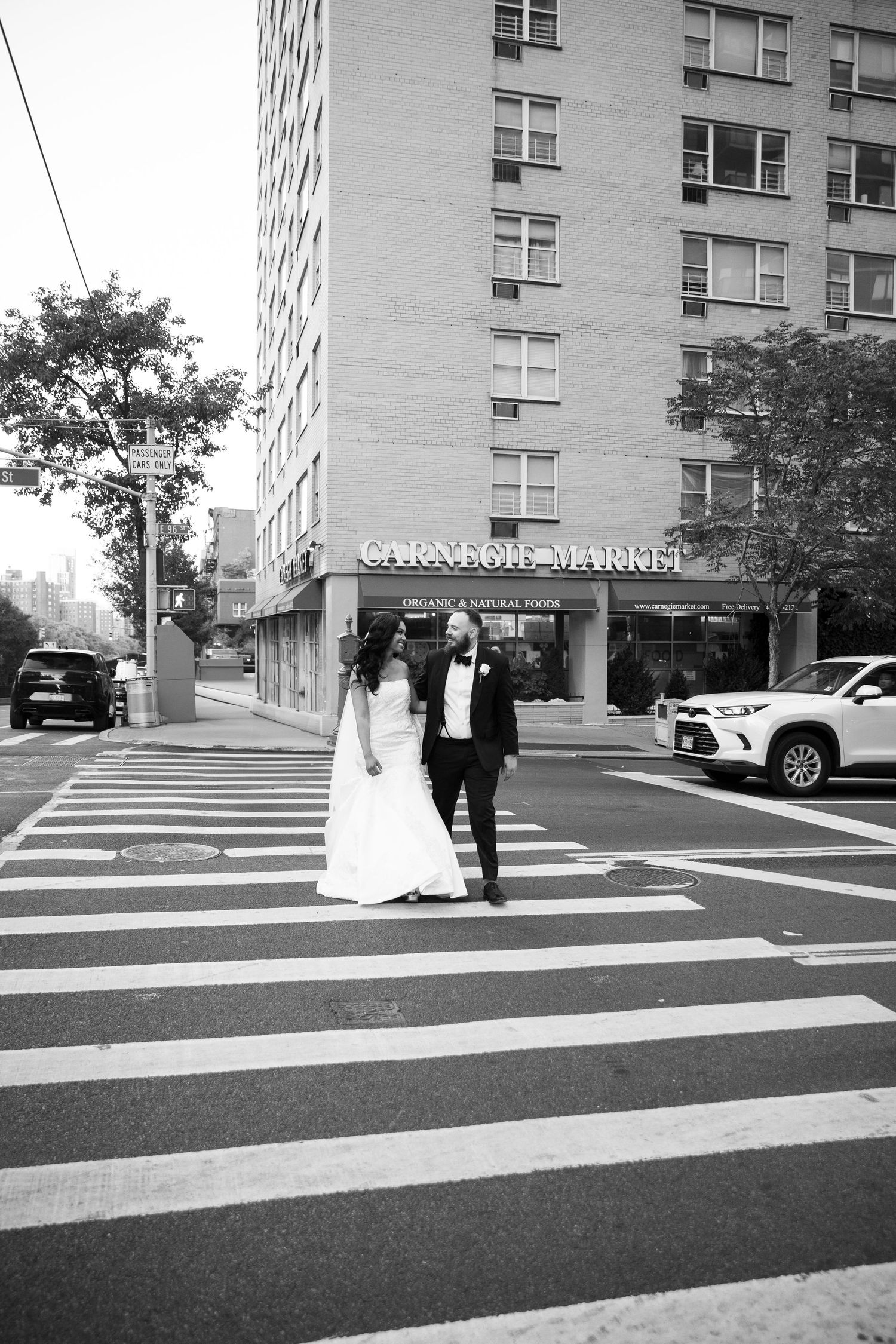 Black and white photo of a couple walking across striped crosswalk near apartment building.