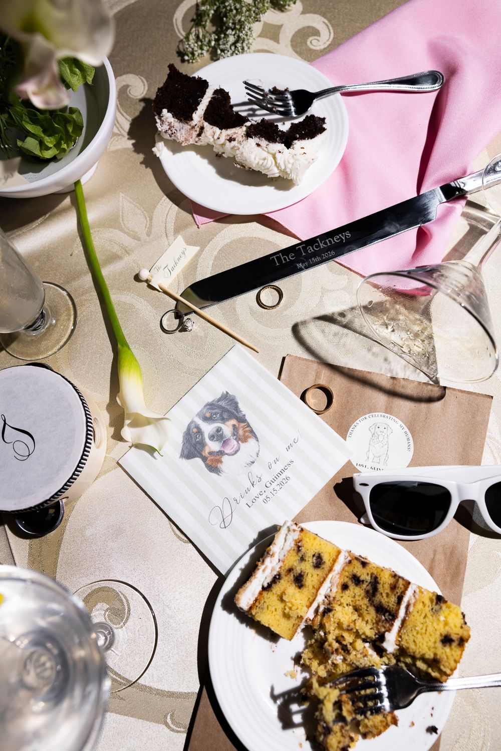 Wedding reception table with cake slices, personalized napkins featuring a dog portrait, sunglasses, and floral decor.