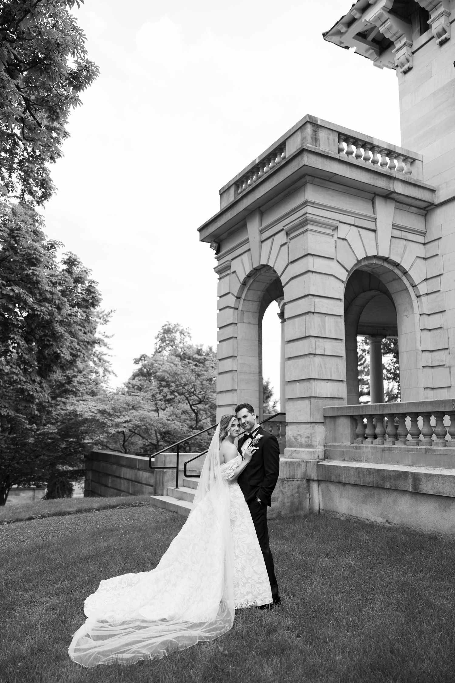 Wedding couple poses outside historic stone architecture with dramatic train flowing on grass.