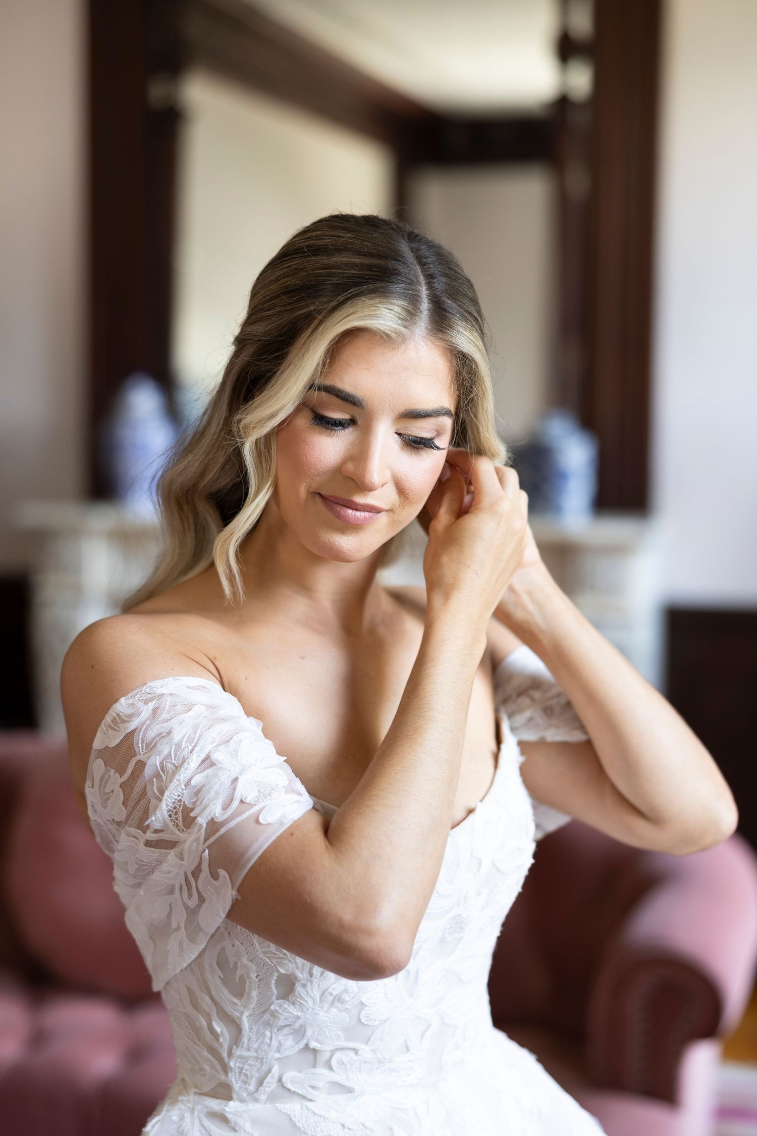 Woman in white lace wedding dress smiles while adjusting her hair in a sunlit room.