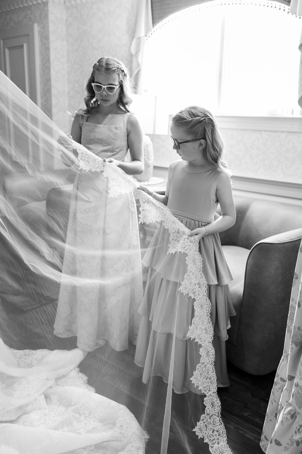 Black and white photo of two girls in dresses examining lace wedding veil indoors.