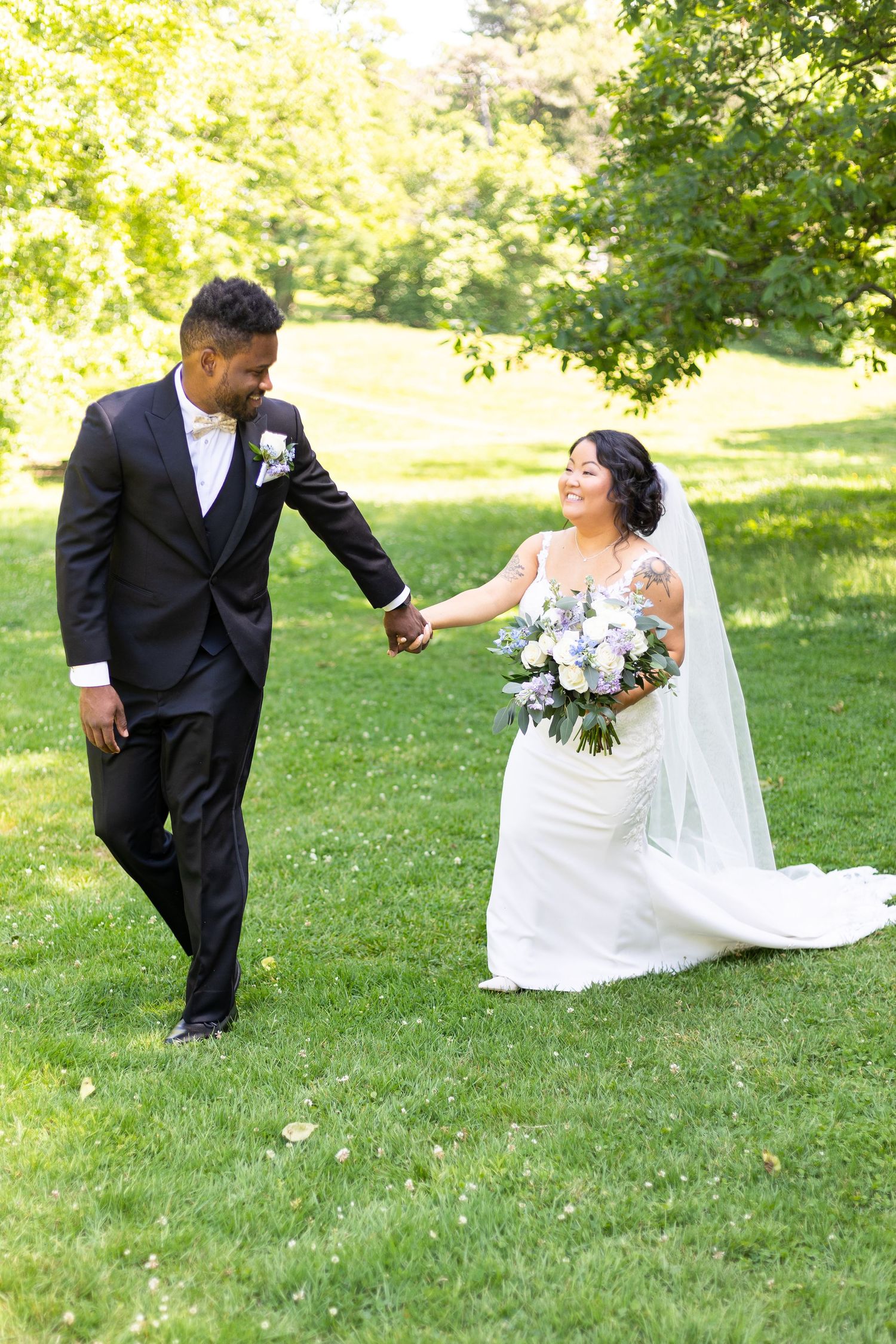 A joyful couple walks hand in hand across a sunlit lawn on their wedding day.