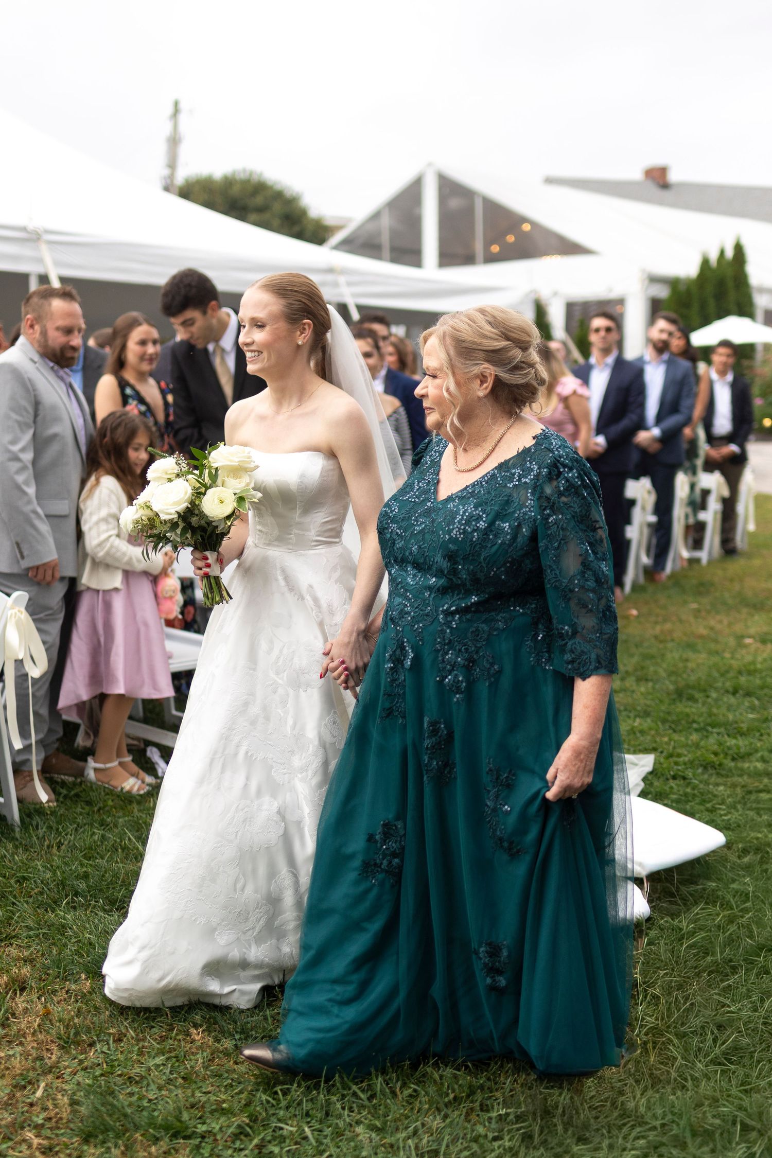 A bride and companion walk down the aisle together at an outdoor wedding ceremony.