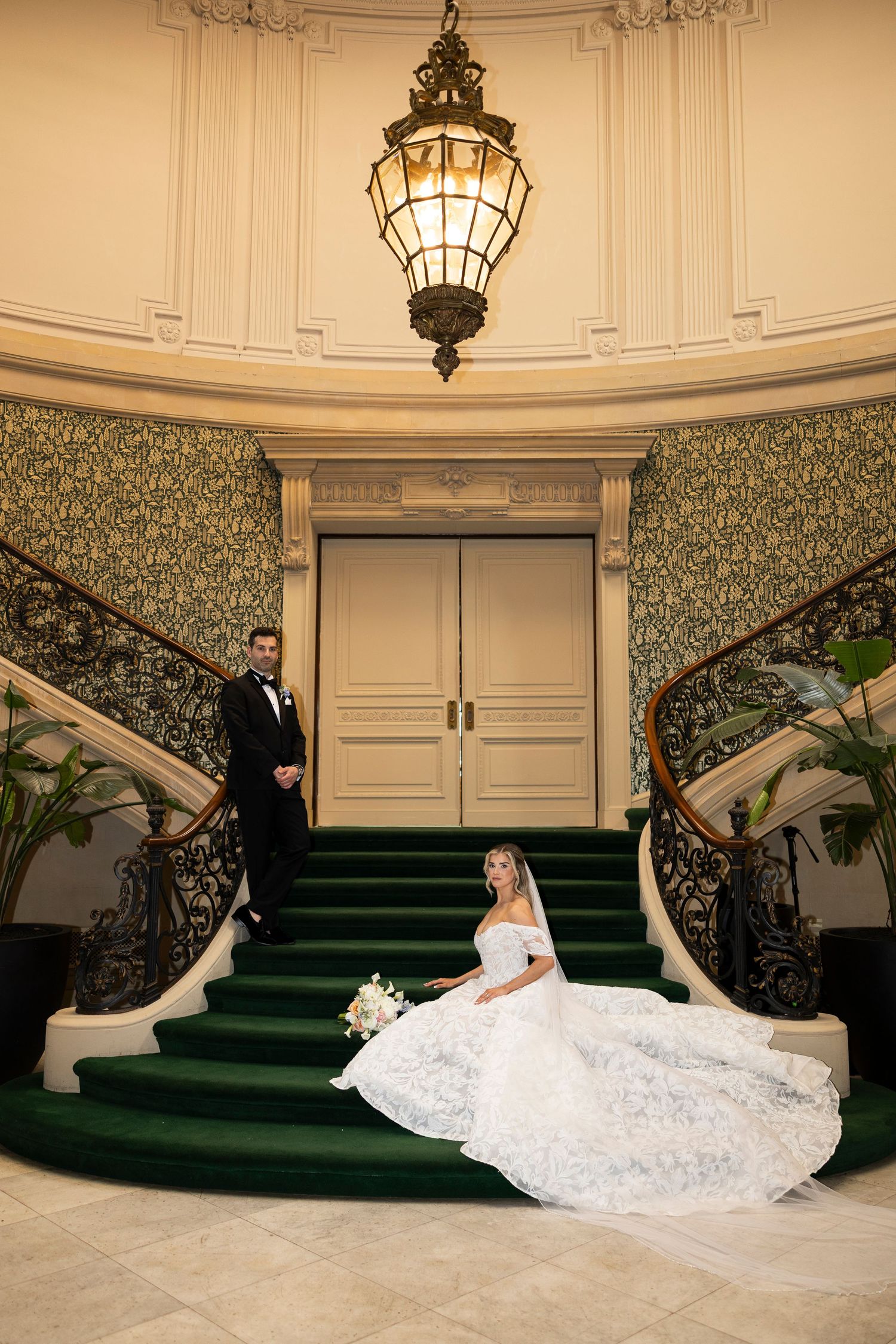 A flowing white wedding dress is dramatically displayed on elegant green stairs in an ornate interior setting.
