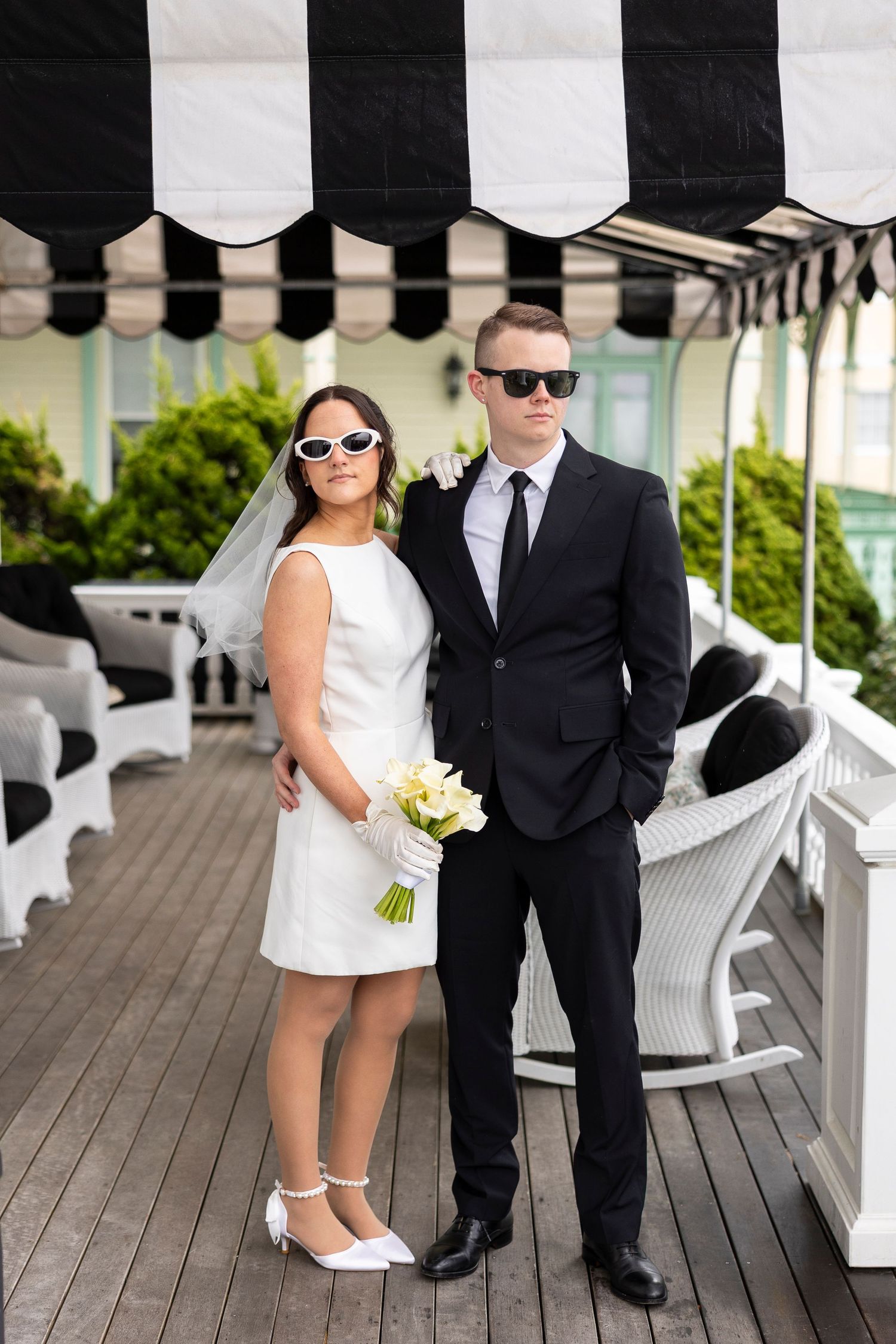 A couple in formal attire poses on a white porch, with the woman wearing a short white dress and sunglasses.
