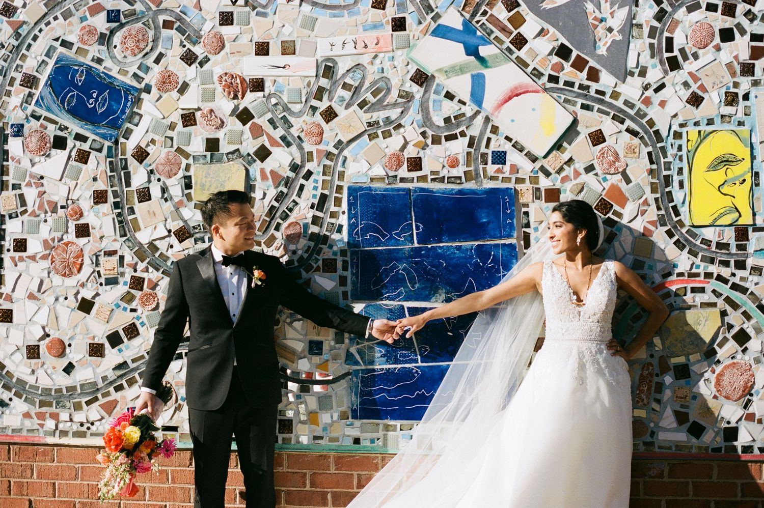 Bride and groom hold hands in front of a colorful mosaic tile wall, bride wearing a white gown with floral bouquet.