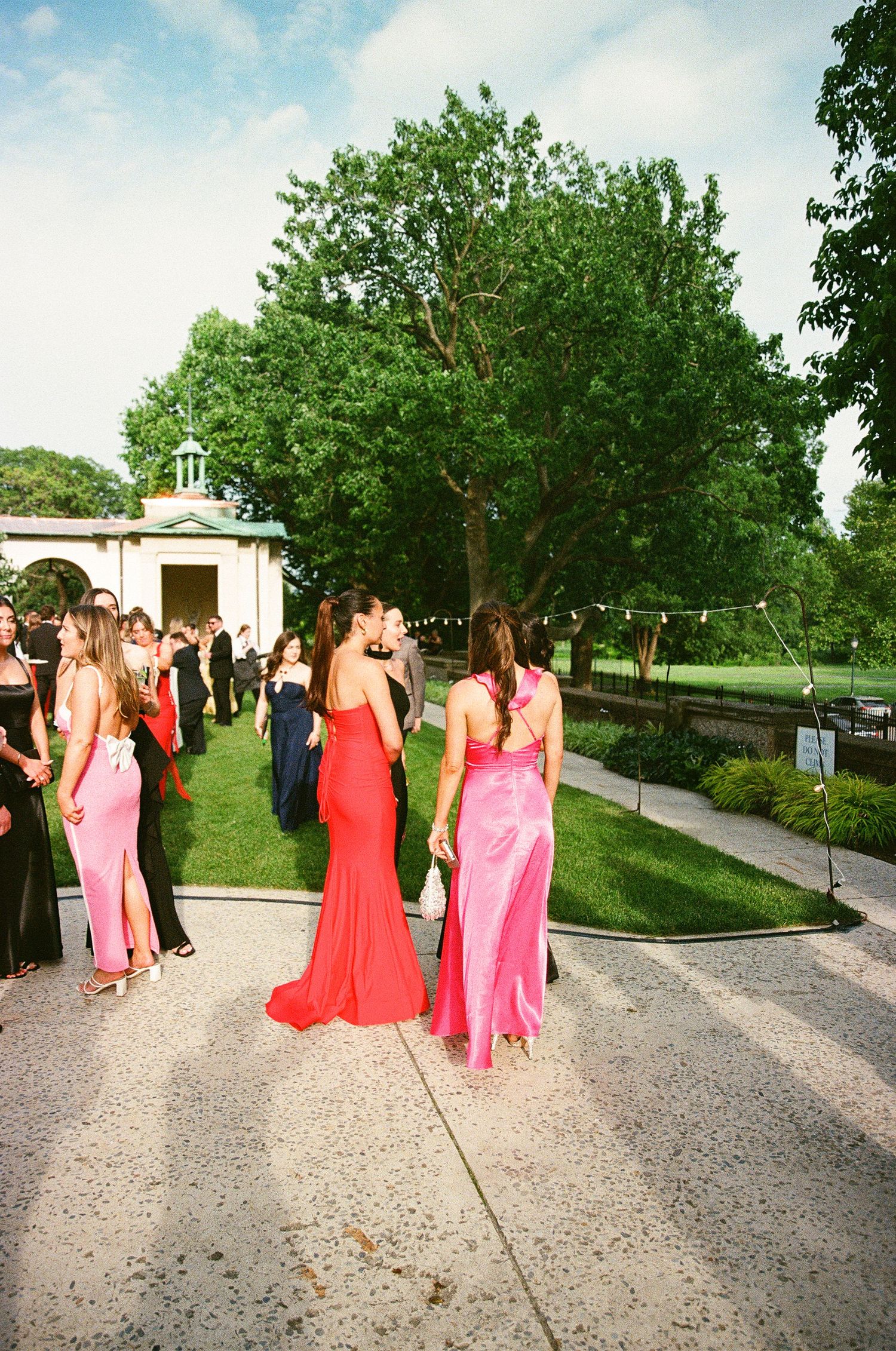 Women in elegant red and pink gowns gather outdoors at a formal garden event with lush green trees.