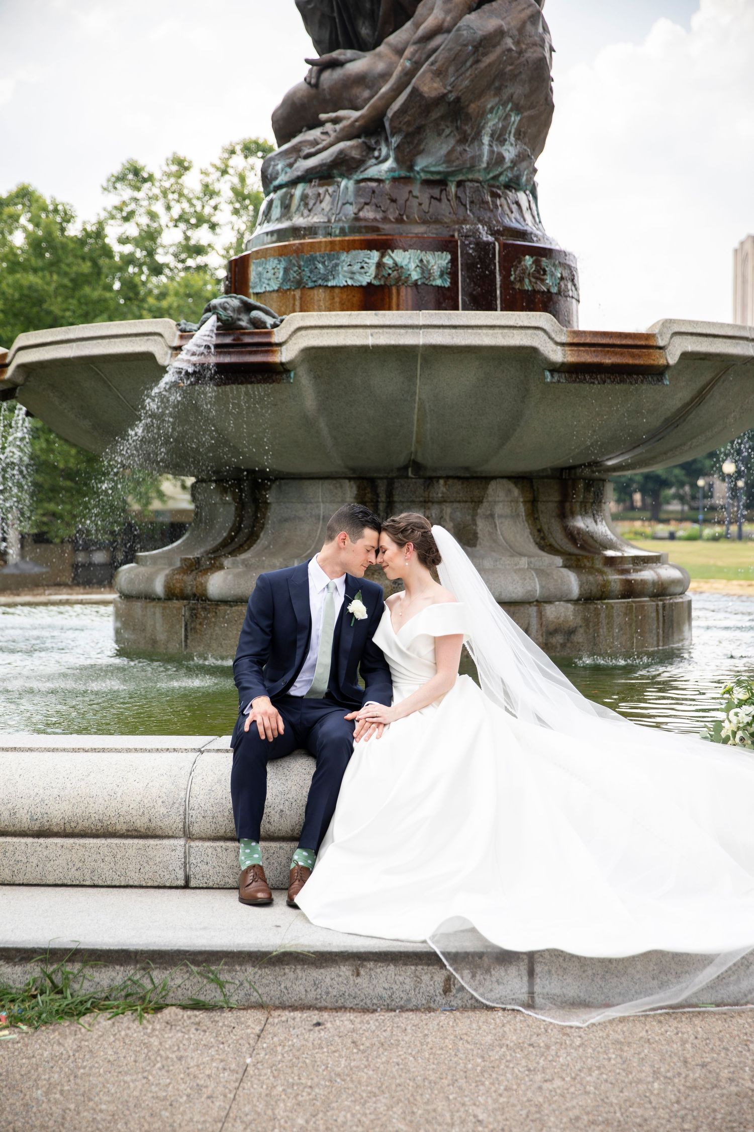 Romantic wedding couple sits by ornate water fountain during outdoor photo session.