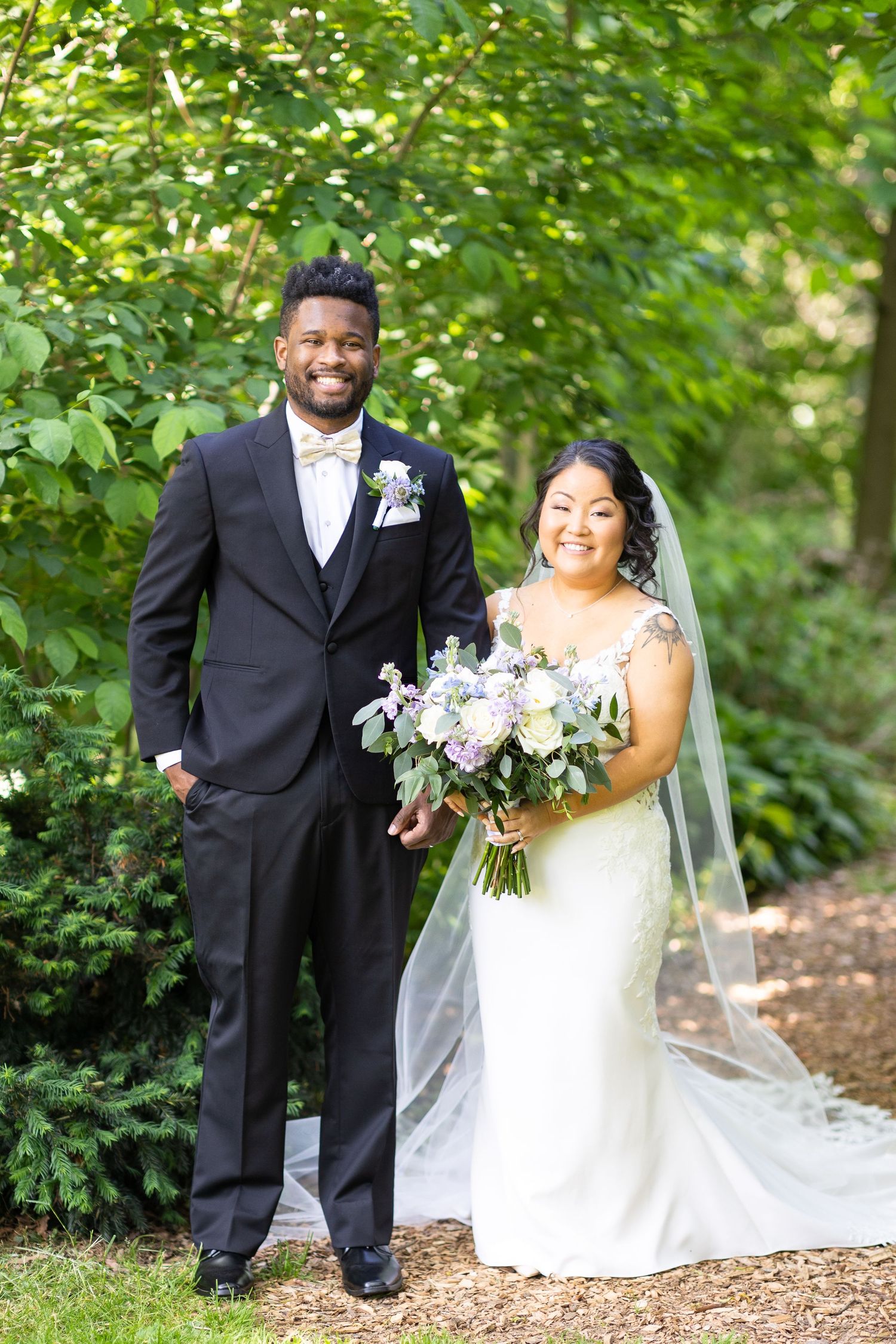 A newlywed couple stands together outdoors holding a purple and white floral bouquet.