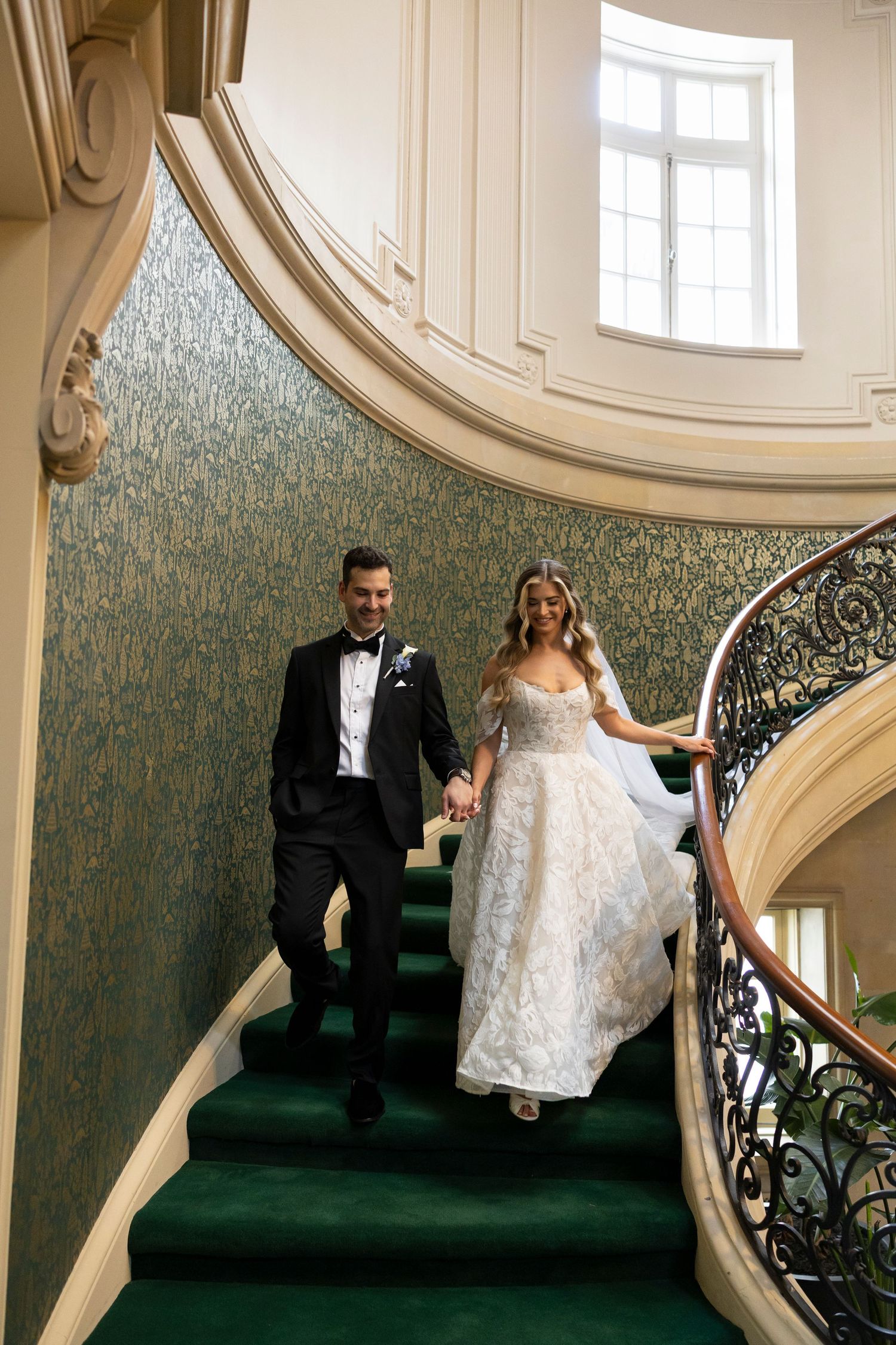 Wedding couple interacts on grand staircase with green carpet and decorative wallpaper.