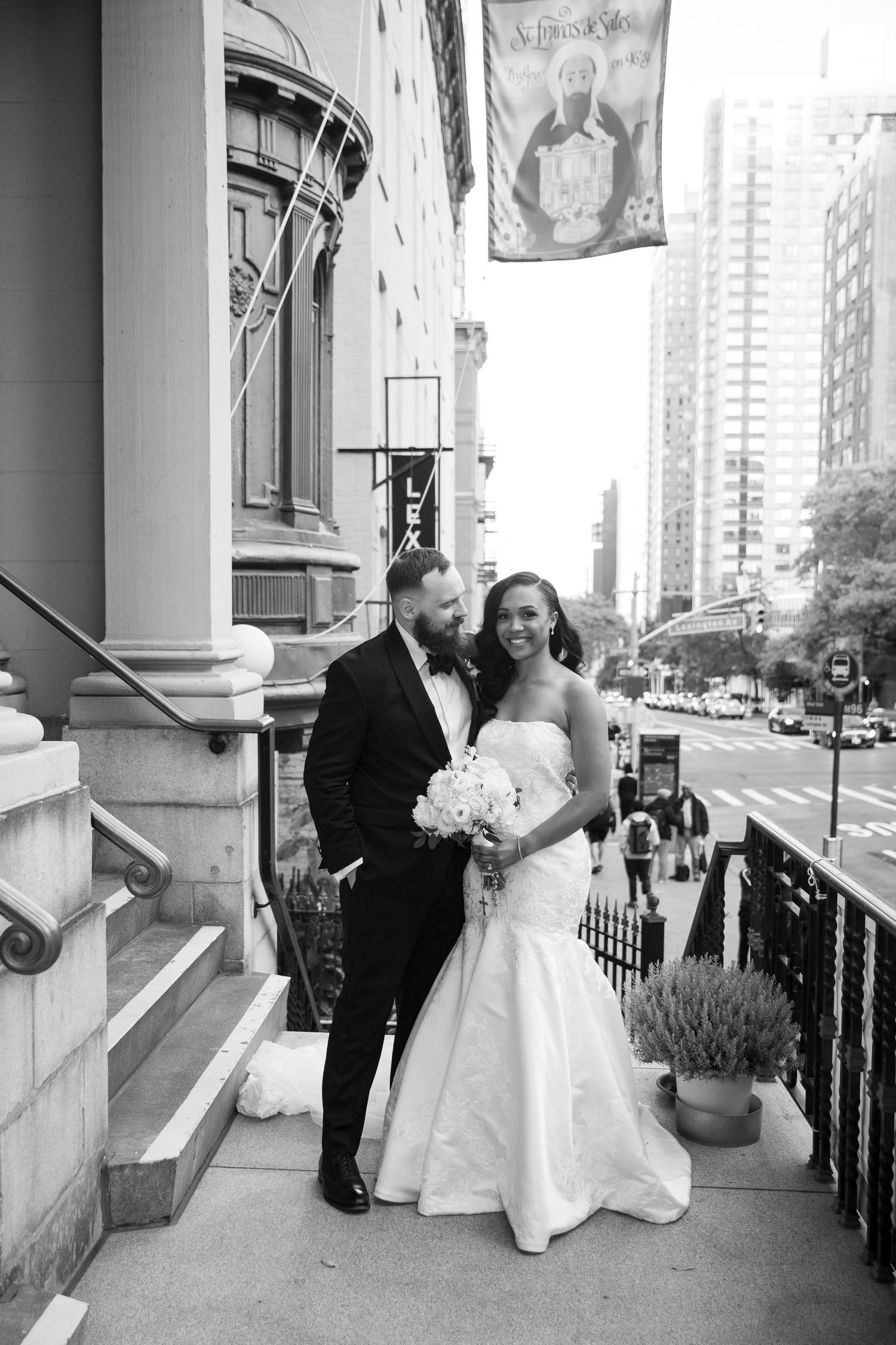 A romantic wedding couple poses on steps outside a city building with white bouquet and formal attire.