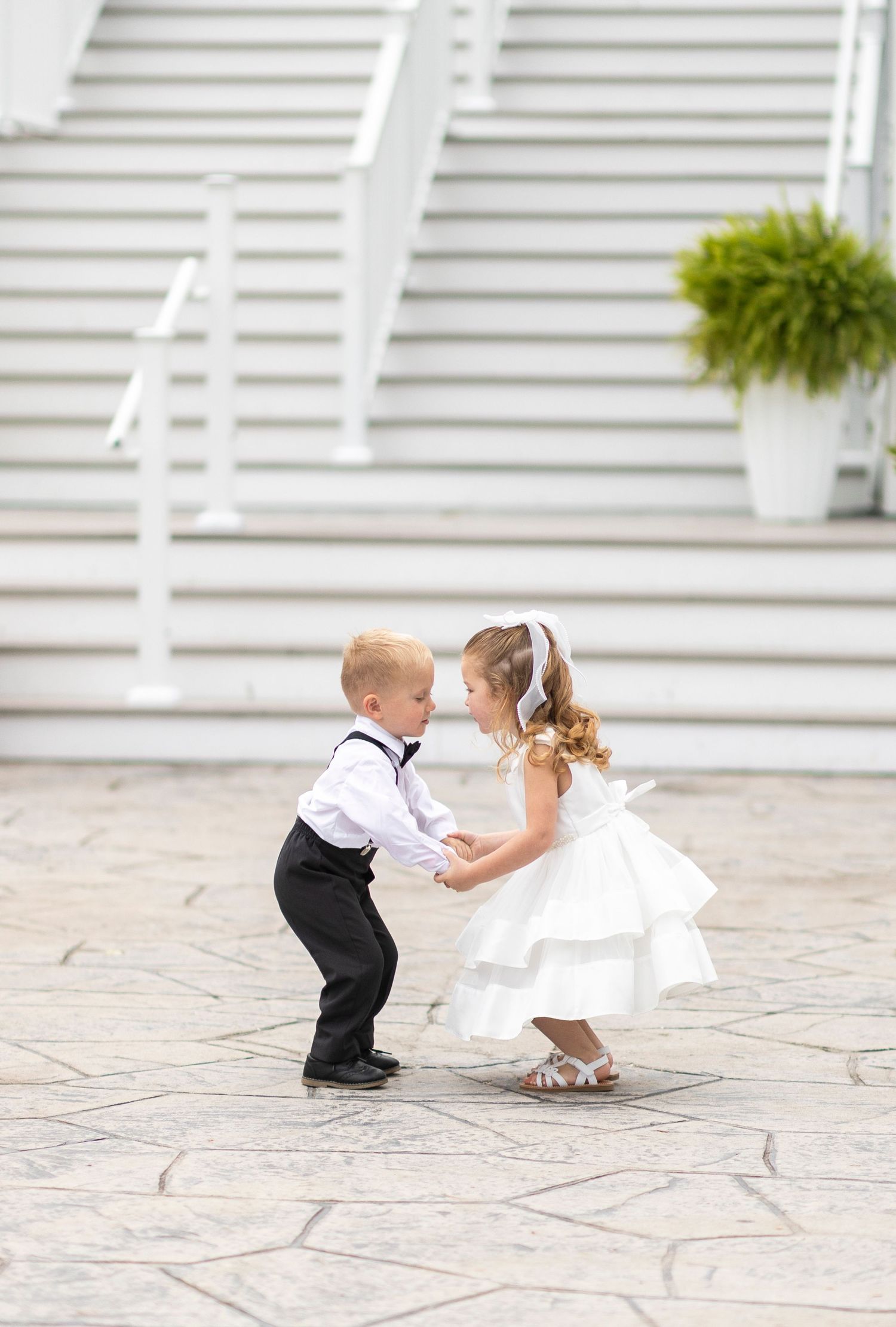 Two young children in formal wedding attire dance together on a stone patio.