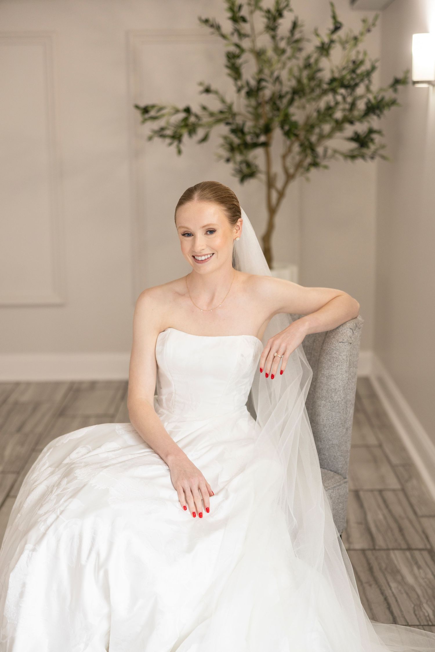 Bride in elegant white wedding dress smiles while sitting on gray couch in light-filled studio setting.