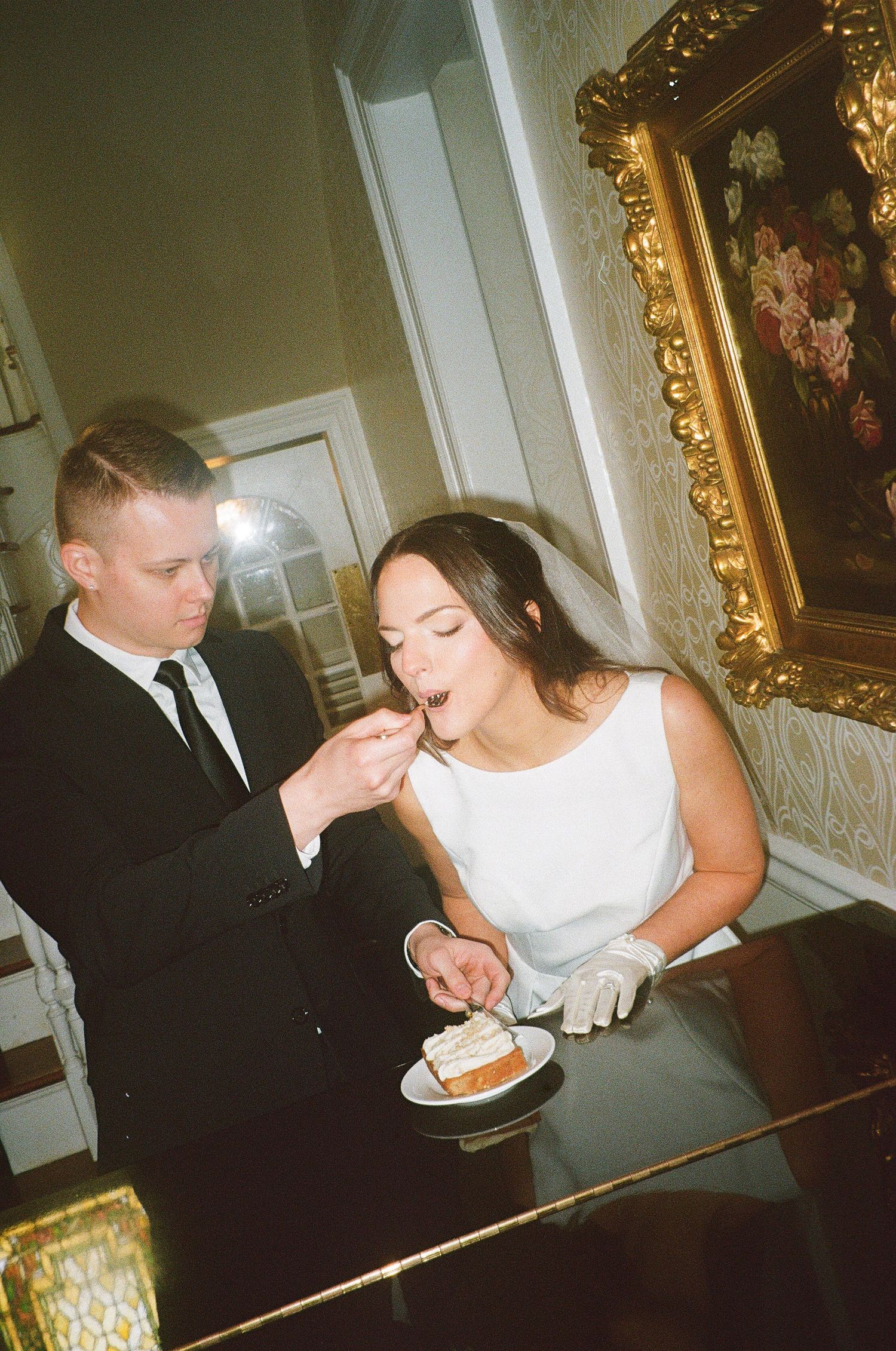 Couple sharing cake at an elegant indoor wedding reception with ornate gold-framed artwork in the background.