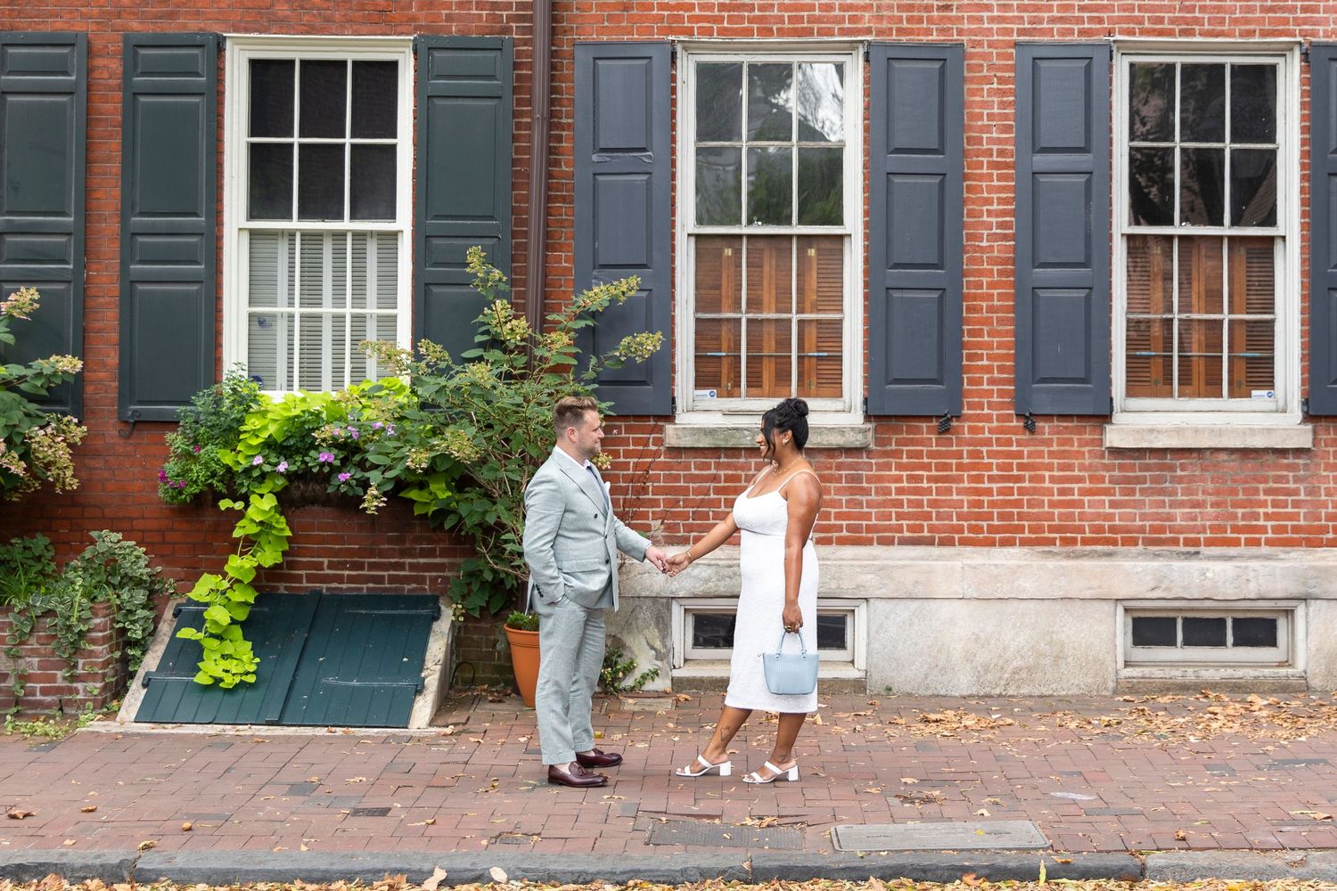 A couple shares a sweet moment on a historic Philadelphia street with red brick buildings and green shutters.