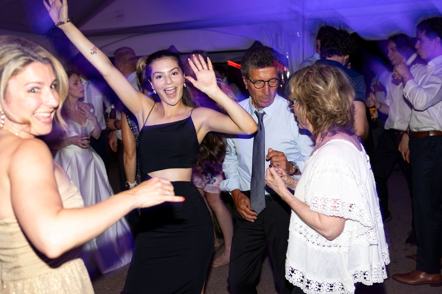 Wedding reception guests dancing with raised arms under purple lighting.