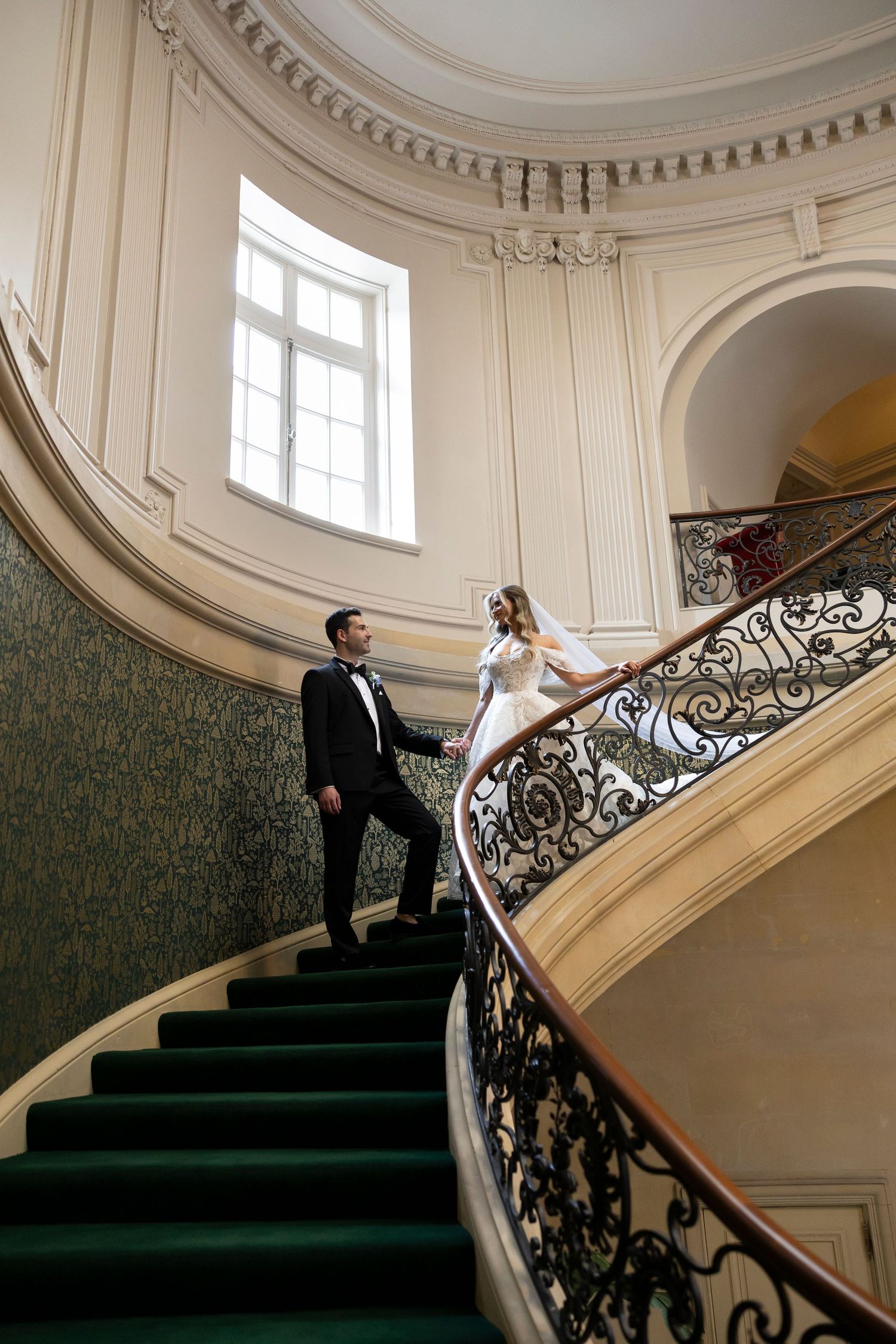 Couple poses on elegant curved staircase with ornate iron railings and green carpet.