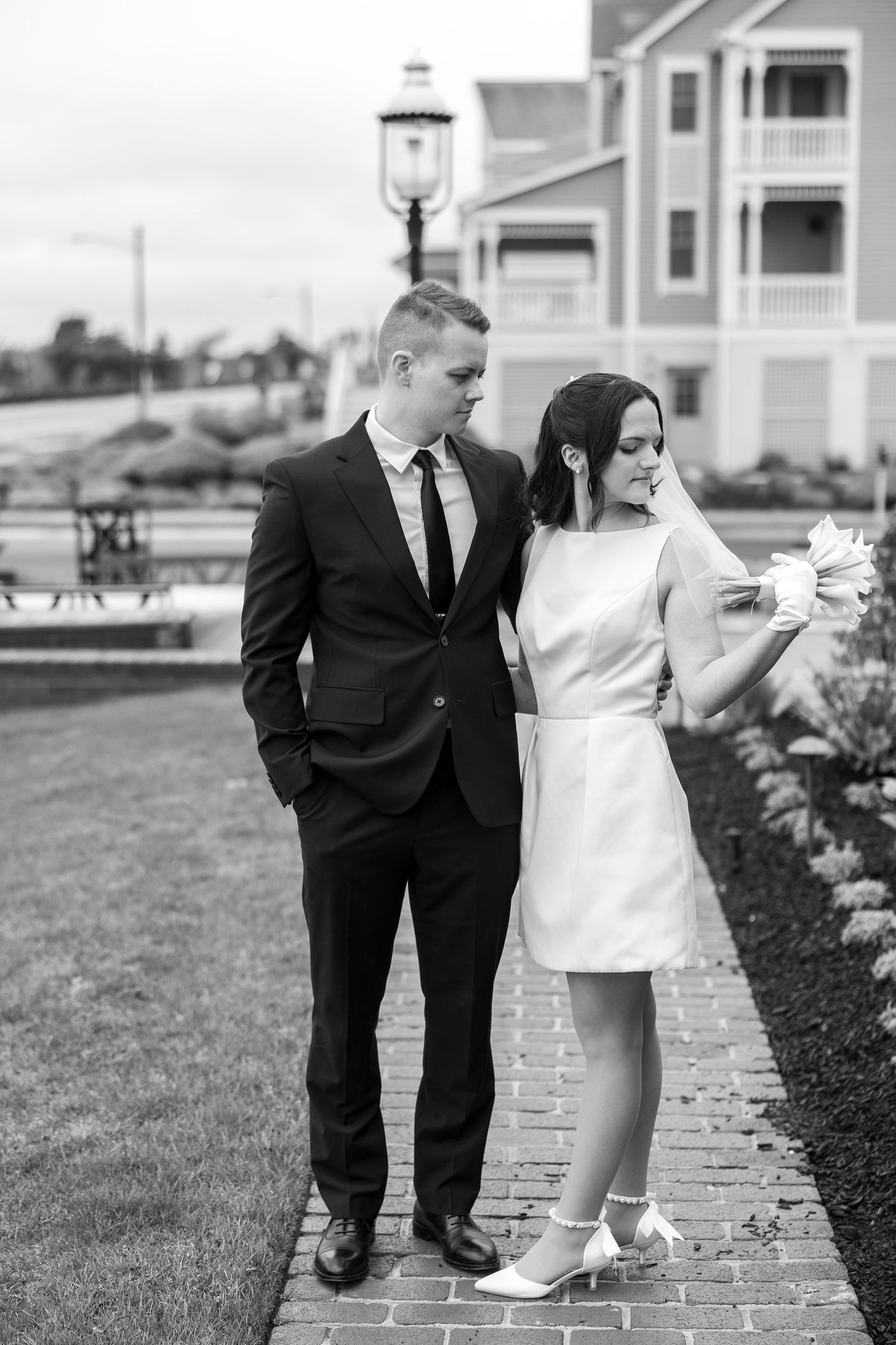 A couple in formal attire poses playfully on a brick pathway outside.