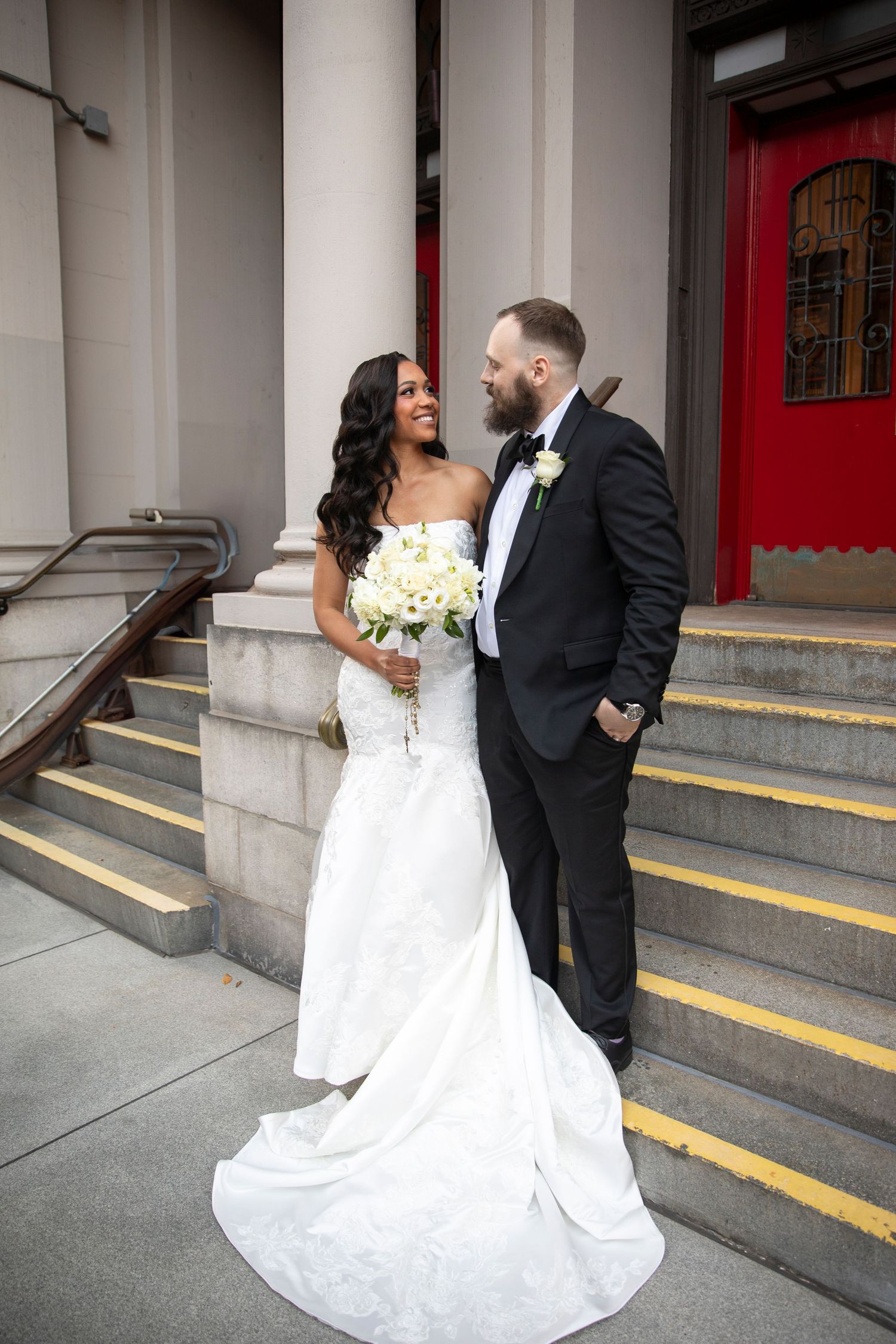 Newlyweds share intimate moments outside a church entrance with red doors and stone steps.