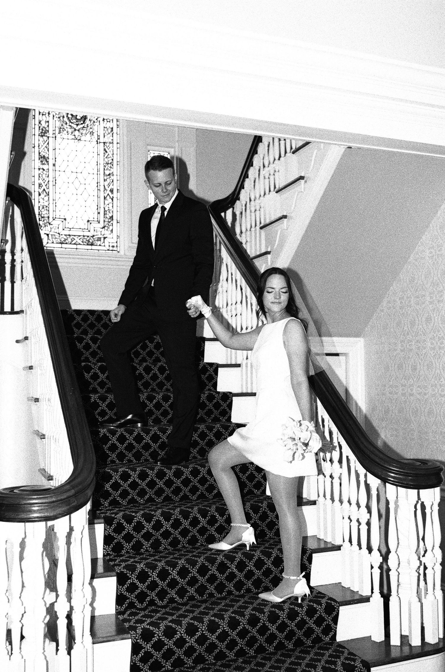 Playful black and white wedding photos showing couple sliding down stair banister in formal attire.