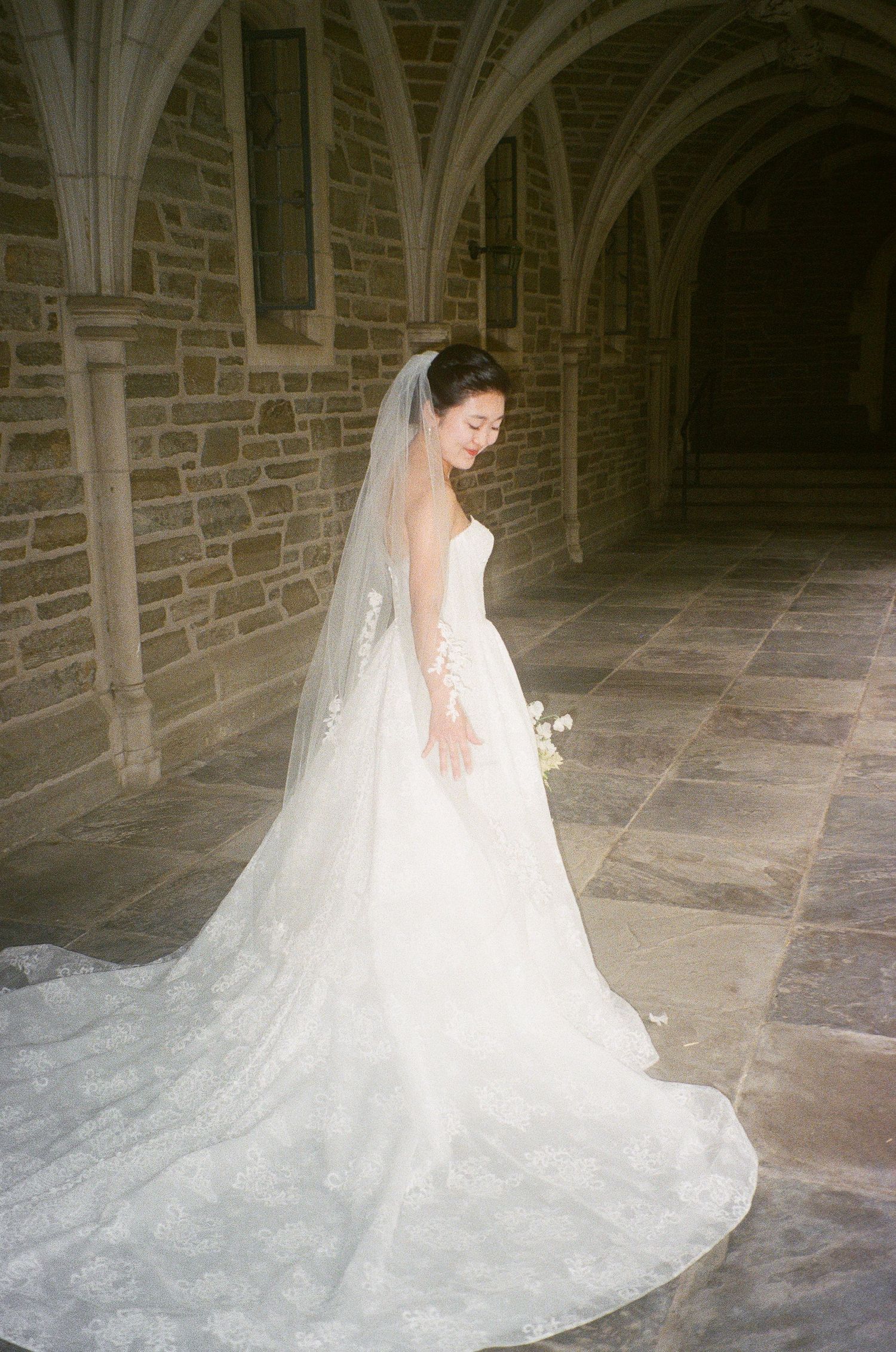 Bride in flowing white gown and veil poses in stone archway corridor.
