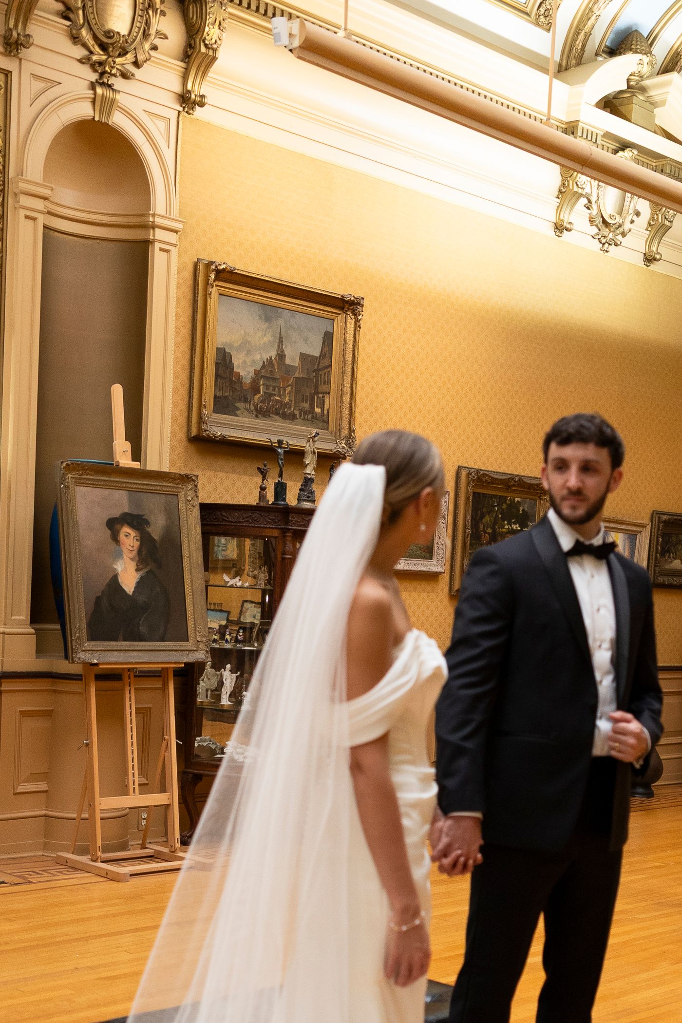 A couple in formal wedding attire dances together in an elegant museum gallery with hardwood floors and ornate walls.