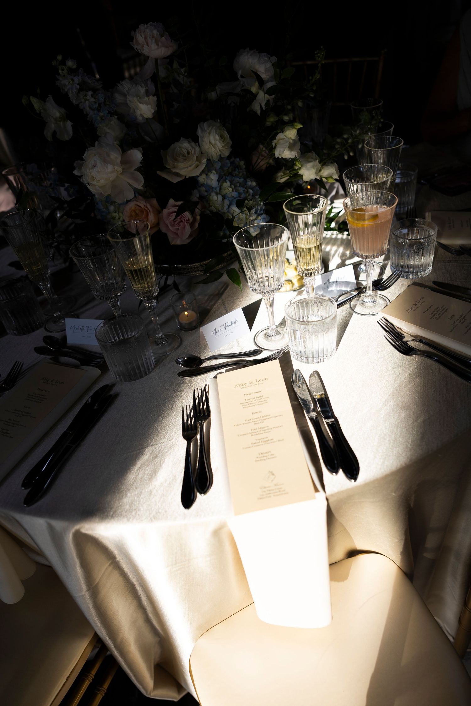 Place settings with menus and glassware illuminated by spotlights on white tablecloths.