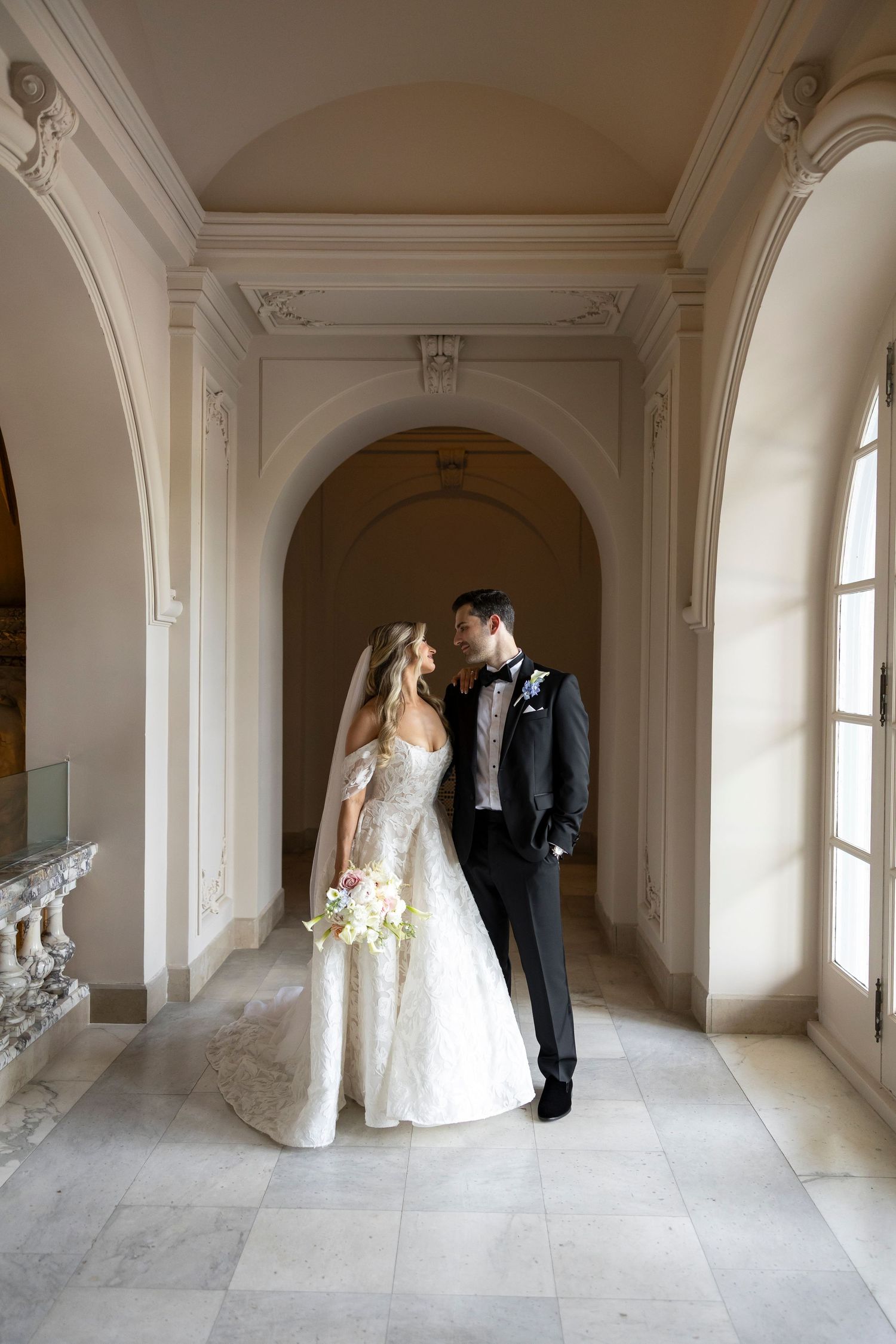 Wedding couple shares intimate moments in an elegant white hallway with arched doorways.