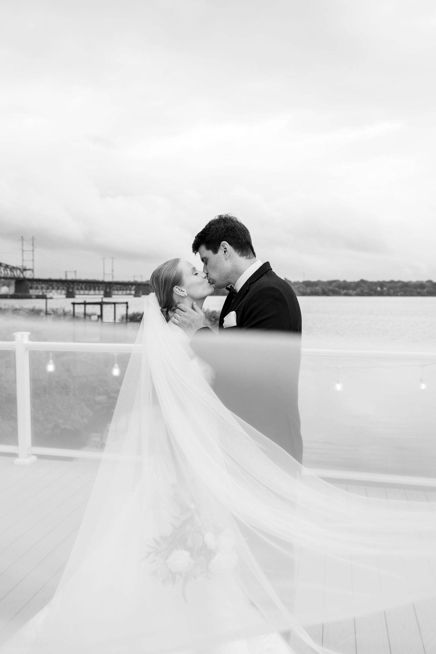 A romantic black and white wedding photo of a couple embracing on a waterfront deck with a flowing veil.