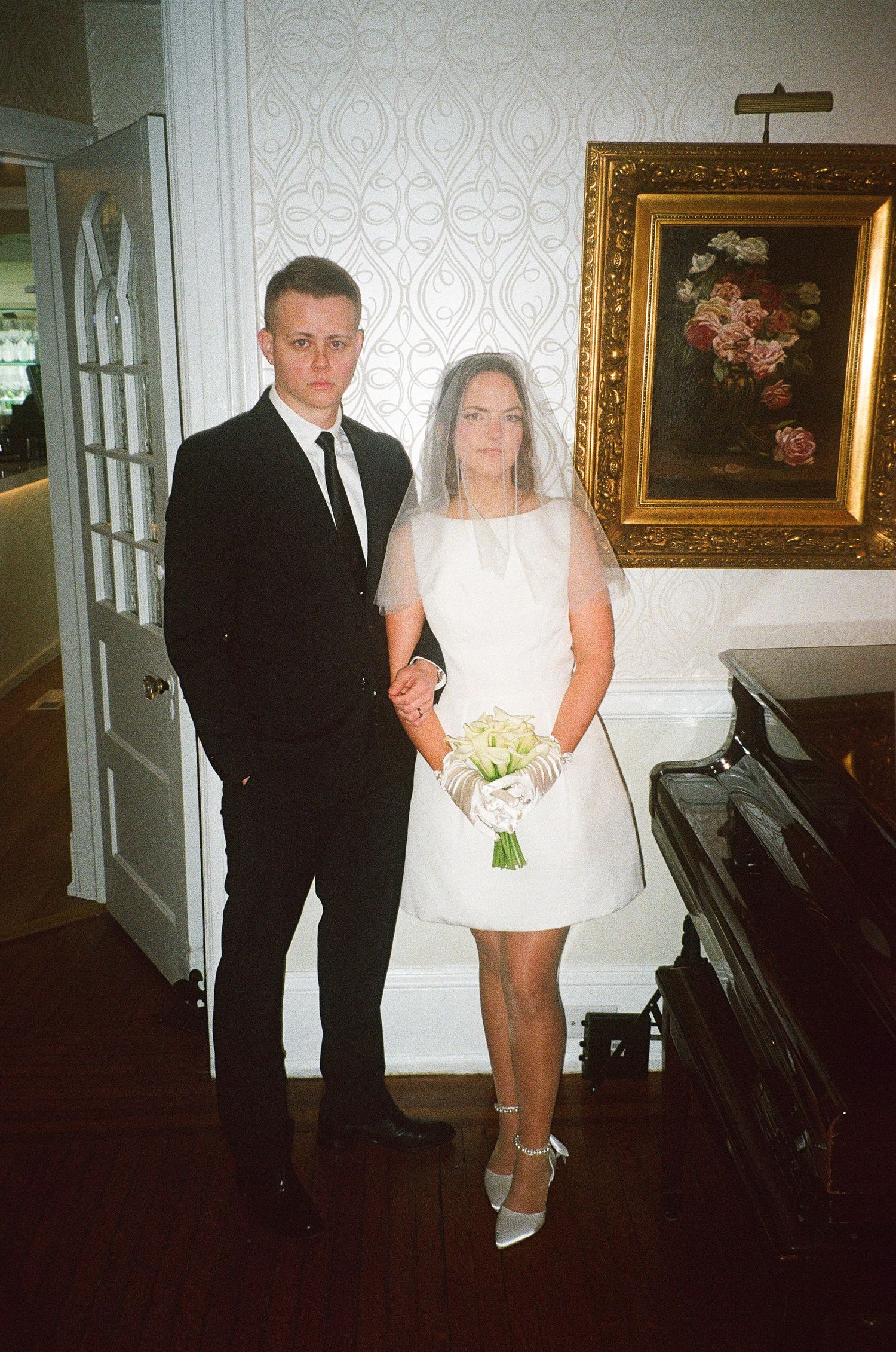 A couple stands together in formal wedding attire near a piano.