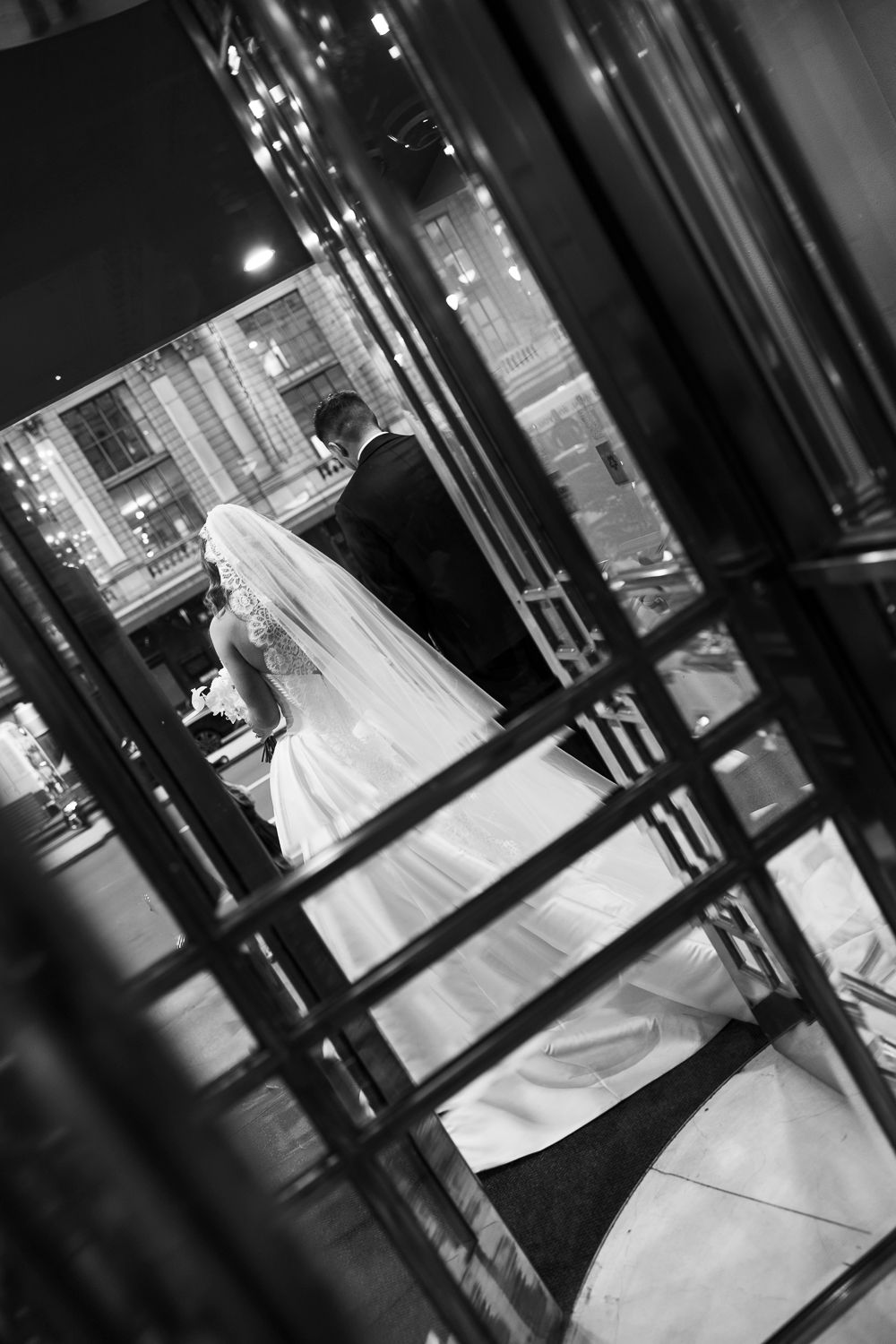 Black and white photo of a bride in a flowing veil and groom walking through elegant venue doors at night.